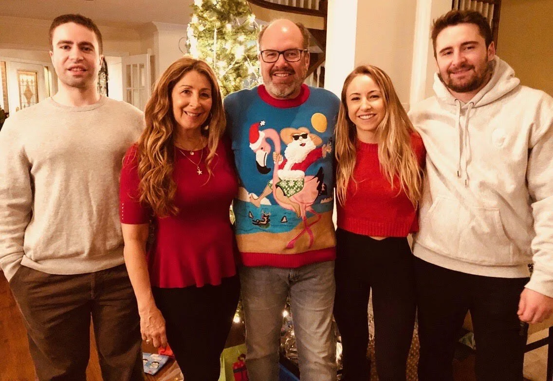 A family of five standing indoors in front of decorated Christmas tree, smiling for the camera. Two men on either end, two women in the middle, and a man with glasses in the center wearing a Christmas sweater.