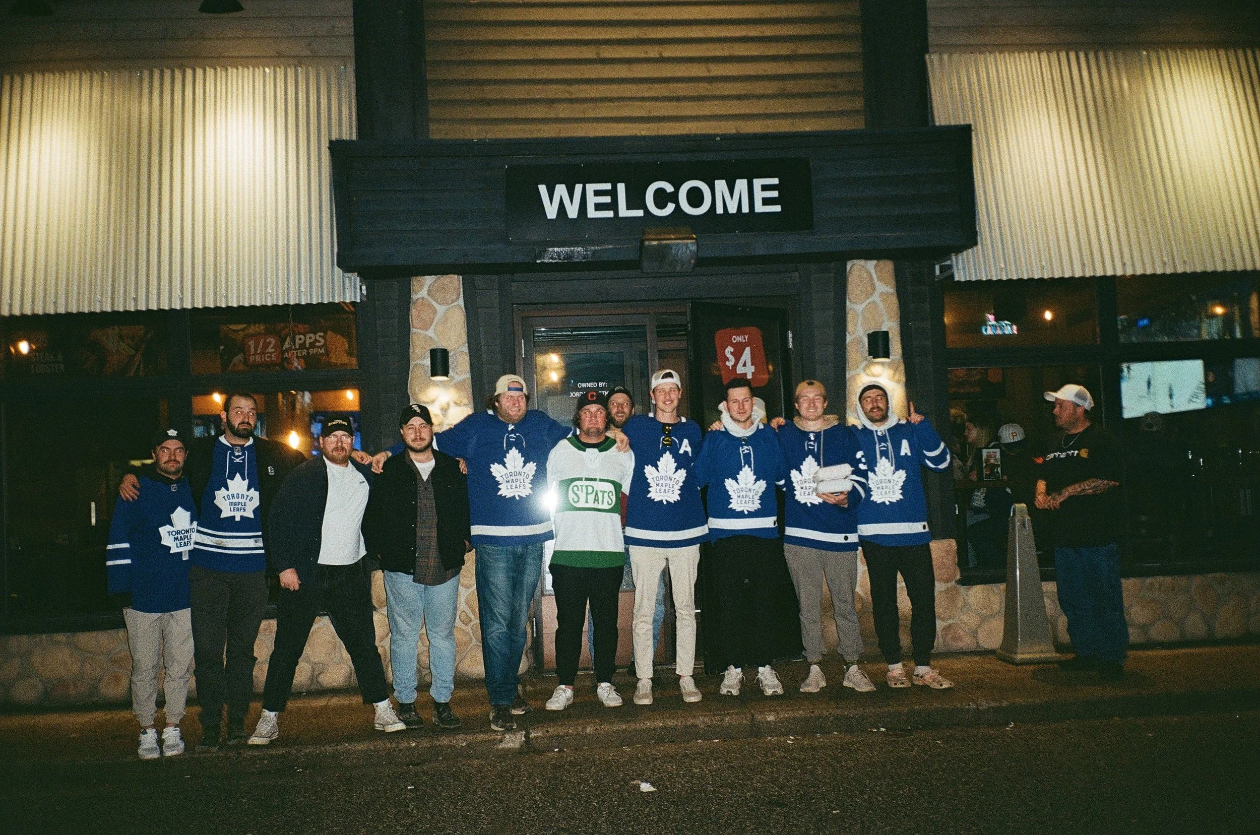 A group of friends standing outside a building at night, with a 'WELCOME' sign above the entrance, some wearing Toronto Maple Leafs jerseys.