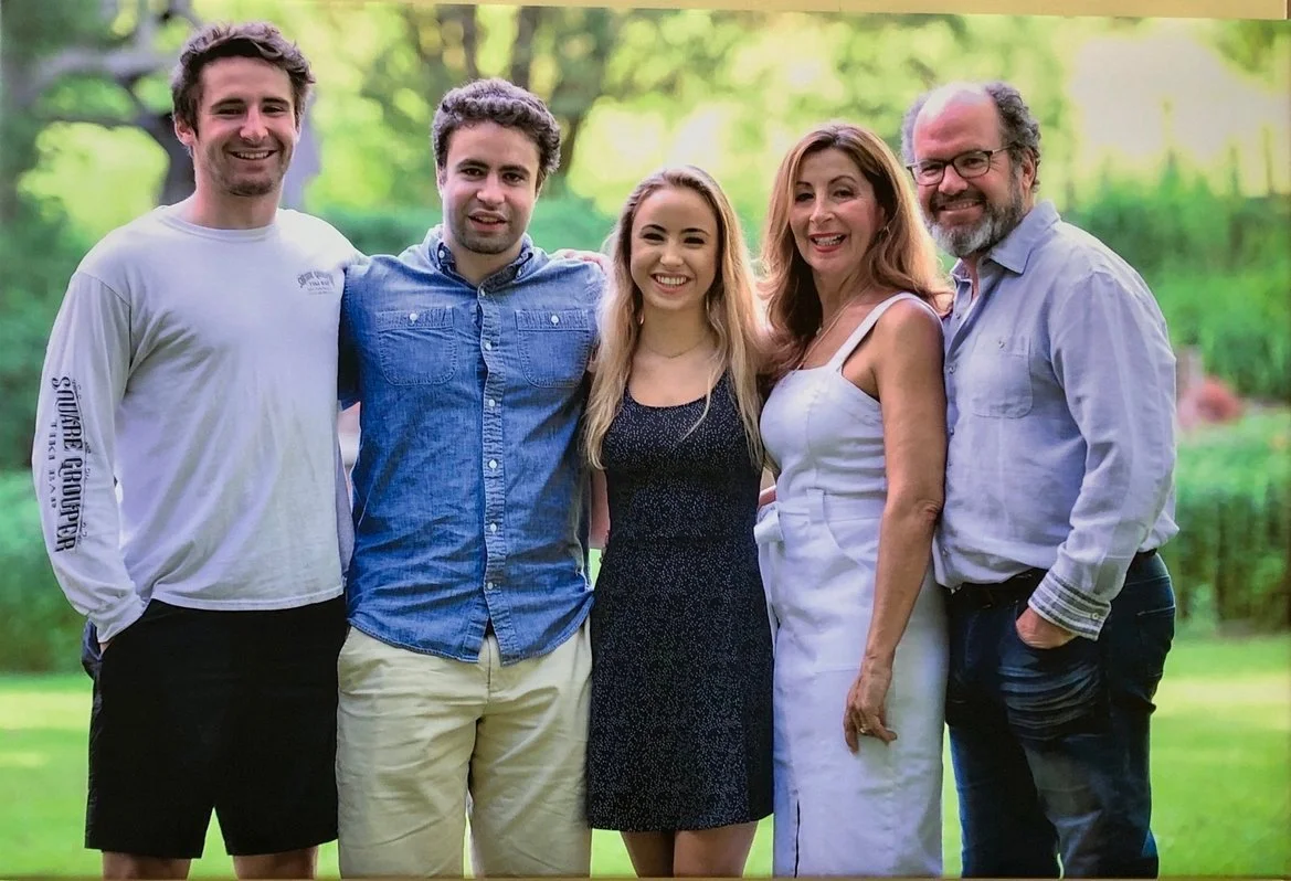 A family of five standing outdoors, smiling, with a lush green background of trees and grass.