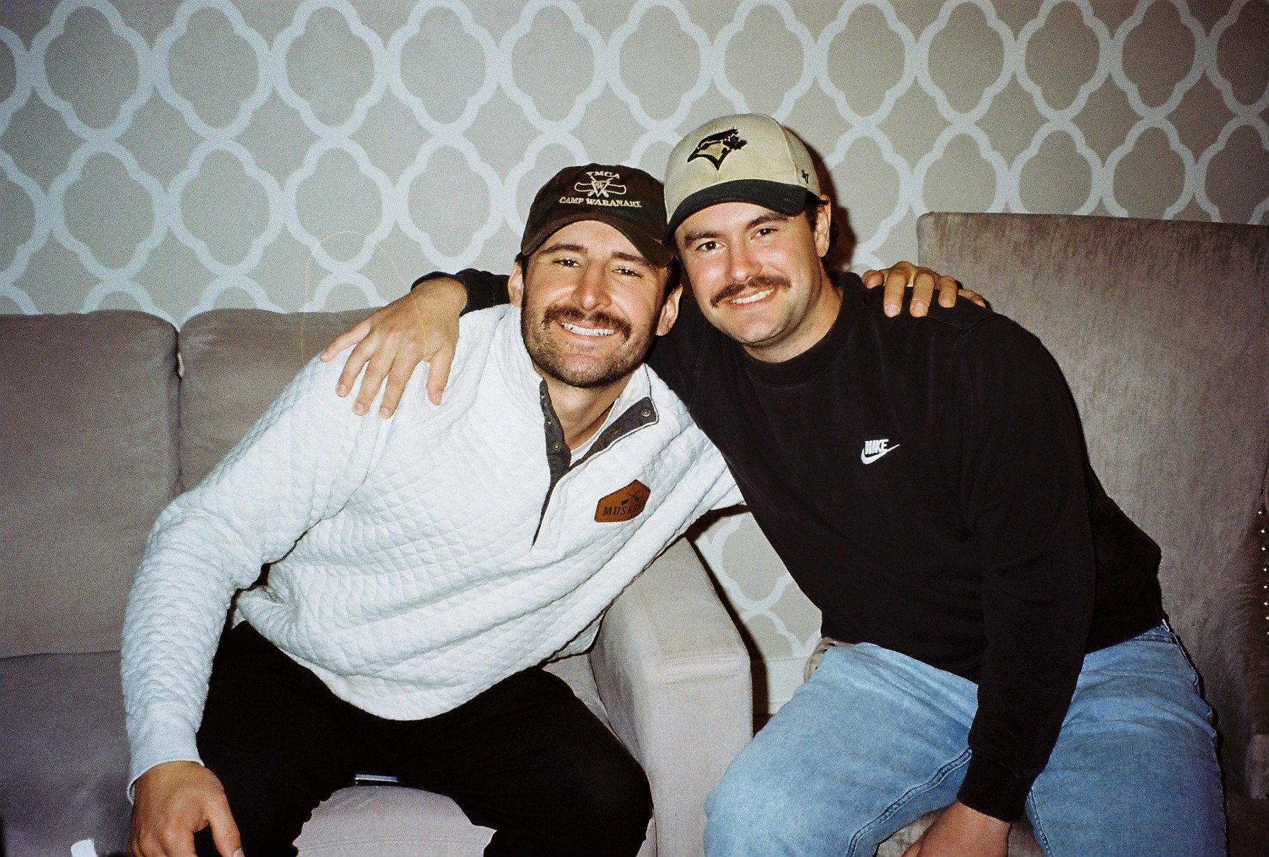 Two men sitting on a sofa, smiling, with arms around each other, in a room with patterned wallpaper.