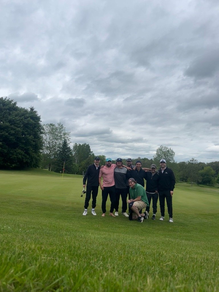 A group of seven men standing together on a golf course fairway with trees and cloudy sky in background, some wearing golf caps and casual athletic clothing.
