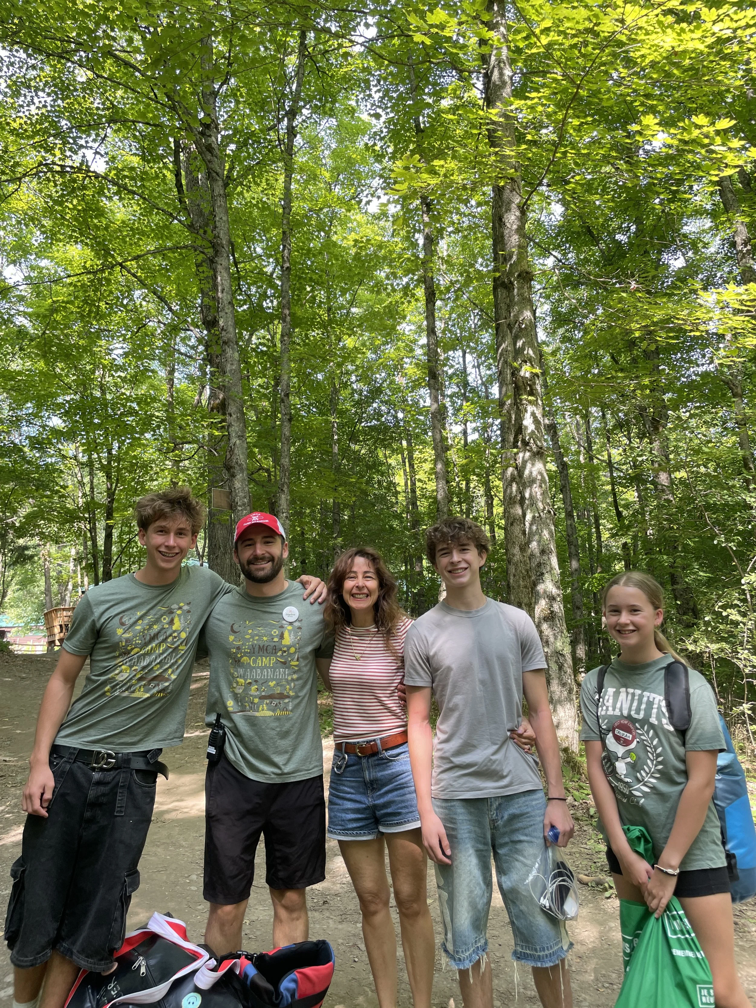 Group of five smiling people standing together outdoors in a forest setting. They are casually dressed, some holding bags or items, with trees and sunlight filtering through leaves in the background.