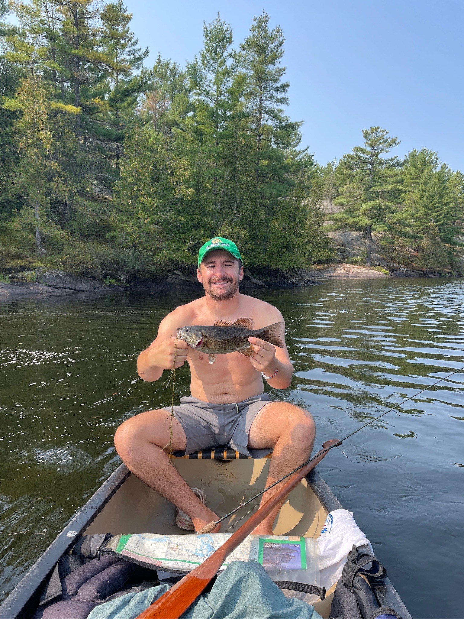 A man sitting in a boat holding a fish, with trees and water in the background, wearing a green cap and smiling.