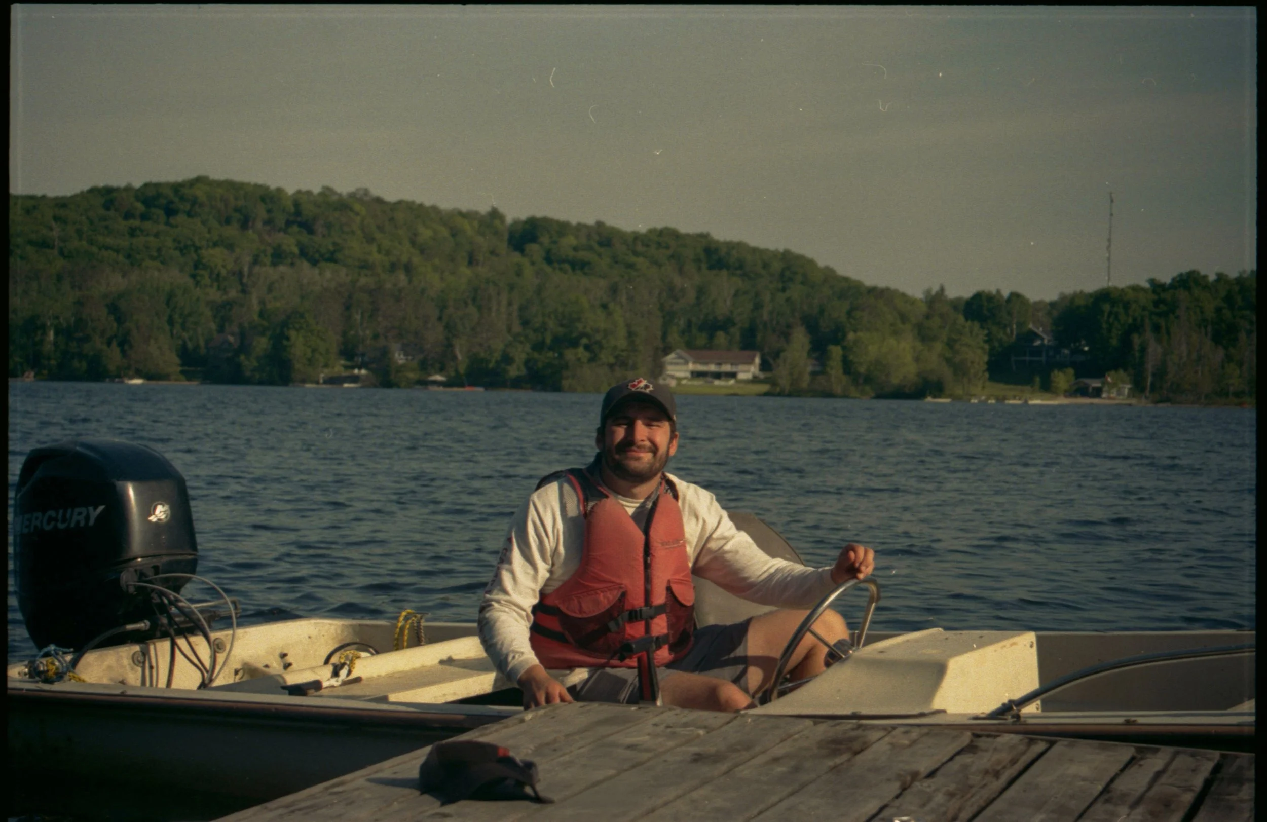 A man sitting in a small boat on a lake, wearing a life jacket and a baseball cap, with a forested hillside and houses in the background.