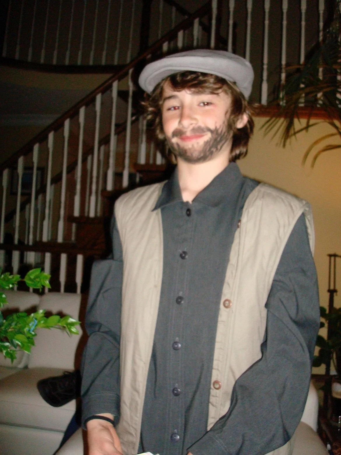 A young man with curly hair and a beard, dressed as a character resembling Han Solo from Star Wars, standing inside a house near a staircase and some plants, smiling at the camera.