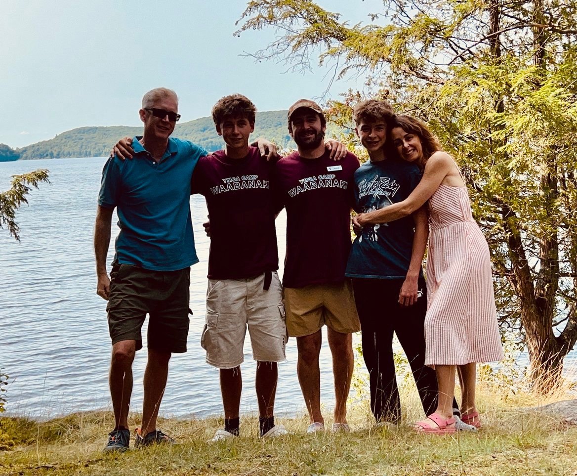 Five people standing outdoors by a lake, smiling and with arms around each other, with trees and hills in the background.