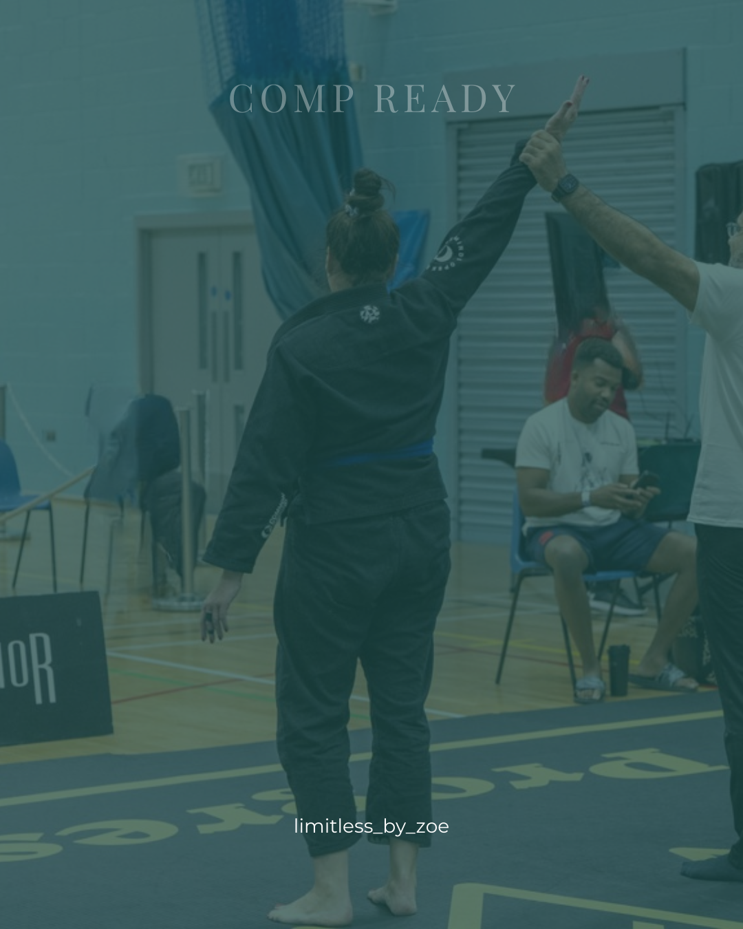 A person with a top bun hairstyle and black jacket is in a gymnasium, raising one arm with two fingers extended, possibly celebrating or signaling. Other people are sitting and walking nearby.