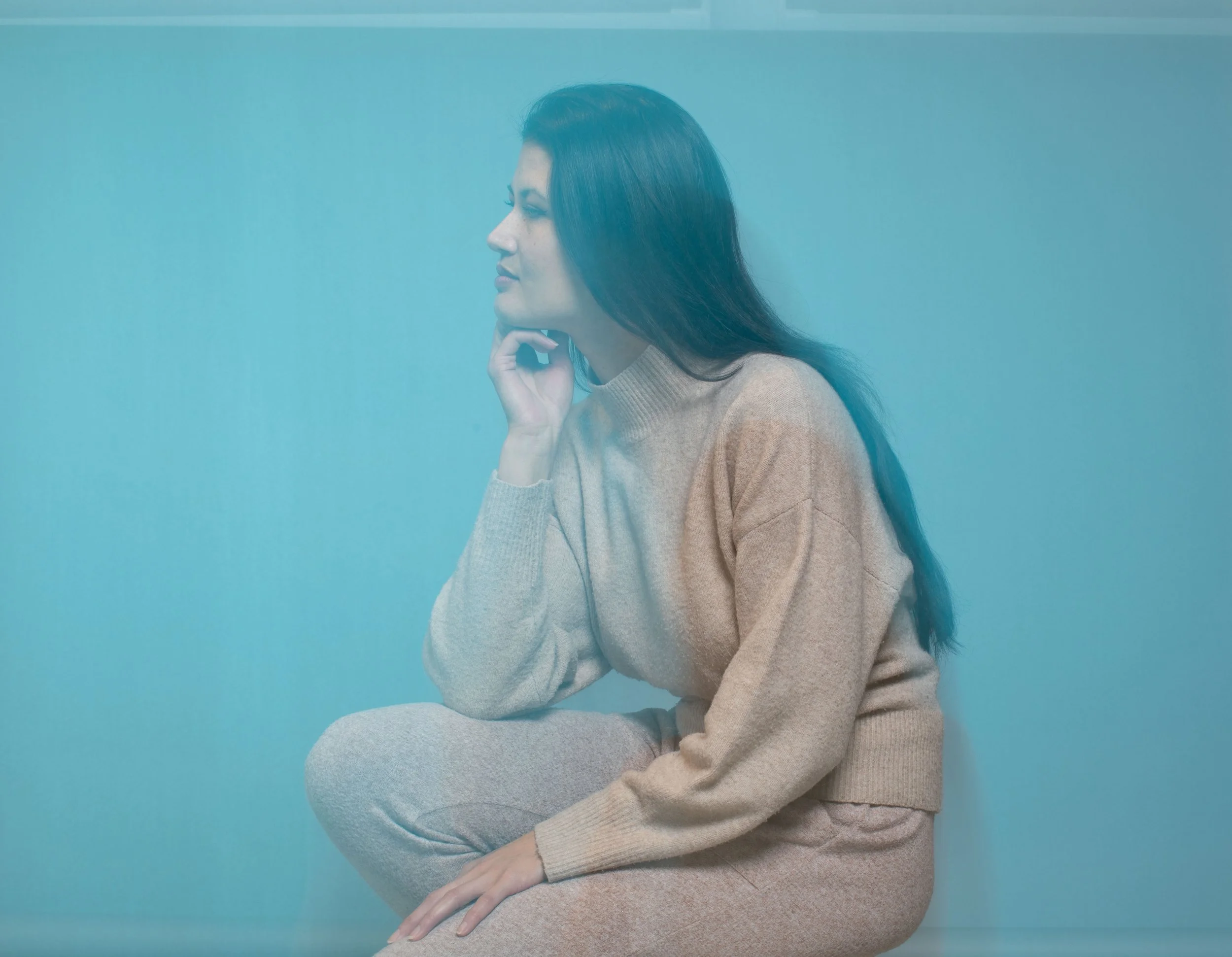 A woman with long dark hair sitting against a blue background, resting her chin on her hand, wearing a beige sweater.