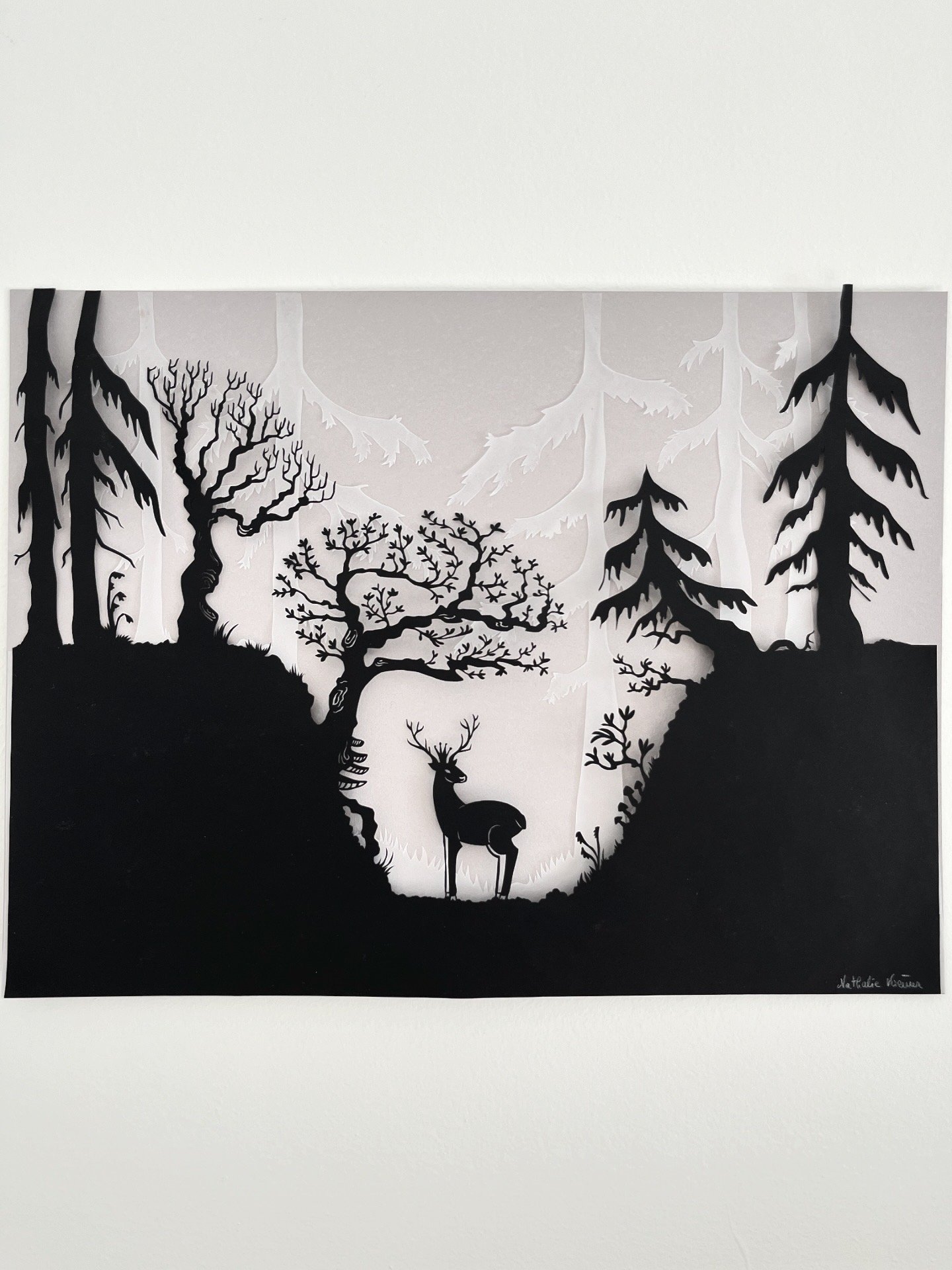 Black and white paper art depicting a forest scene with trees and a deer in the foreground.
