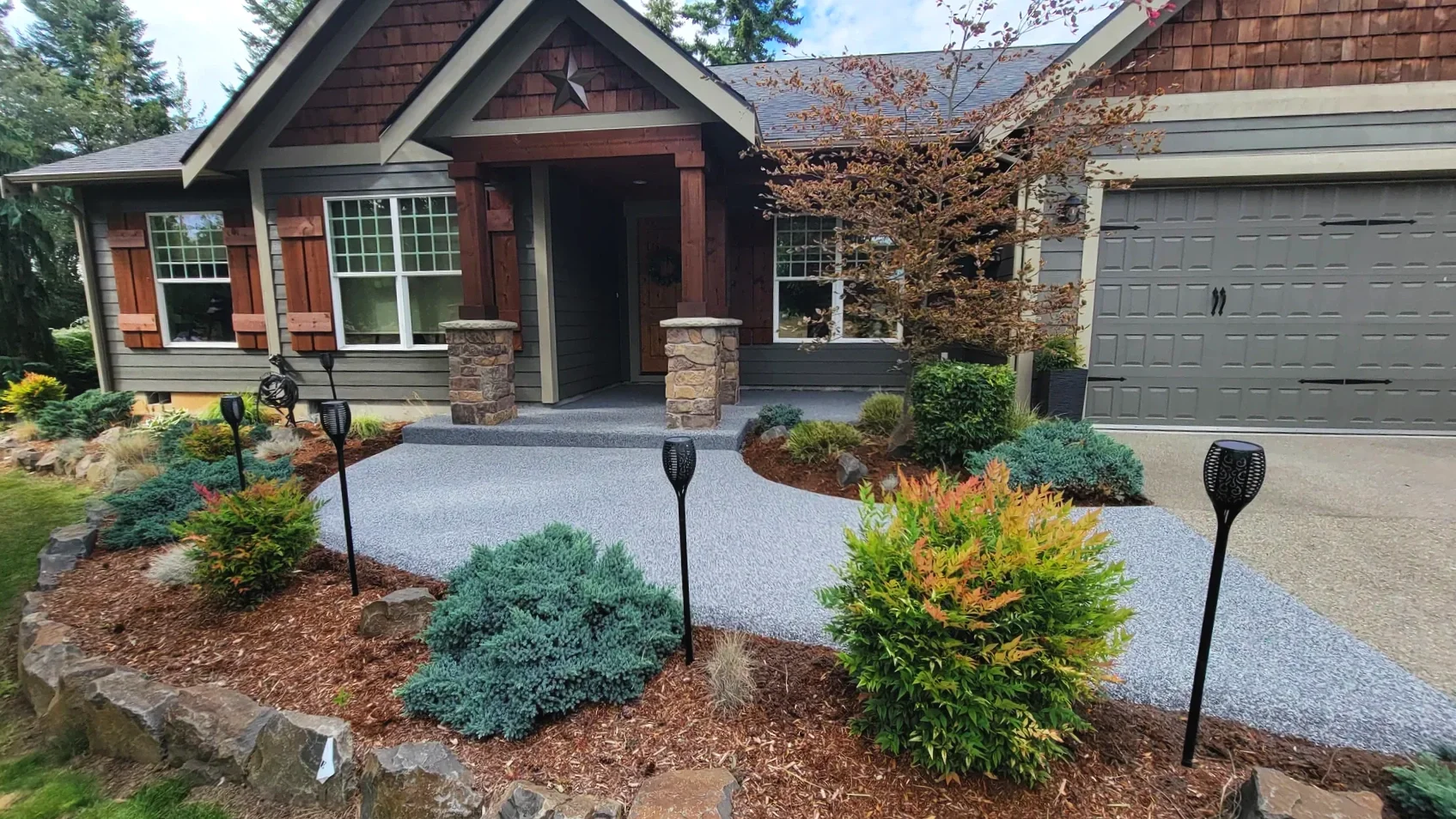 Front view of a house with a stone and wood porch, surrounded by landscaped garden with shrubs, small trees, and solar garden lights.