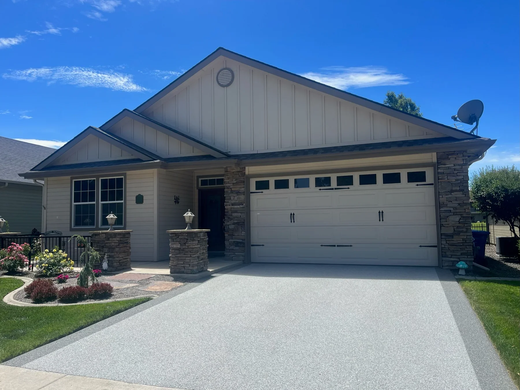 A modern suburban house with a white garage door, stone accents, front porch, and well-maintained front yard with flower beds, surrounded by a concrete driveway and green lawn, under a clear blue sky.
