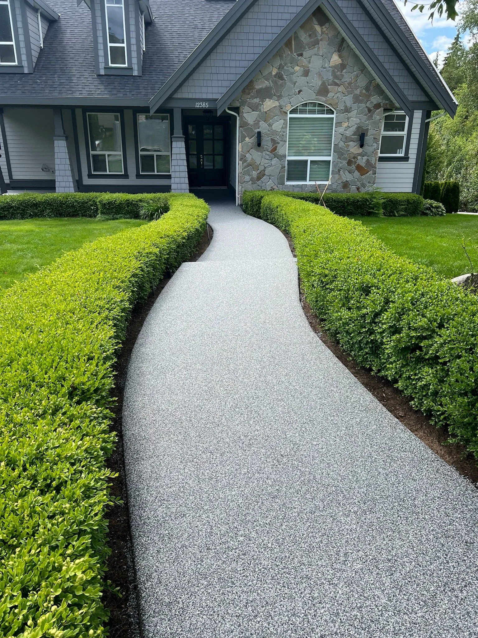 A curved concrete sidewalk leading to the front door of a house, flanked by well-manicured green bushes on both sides.