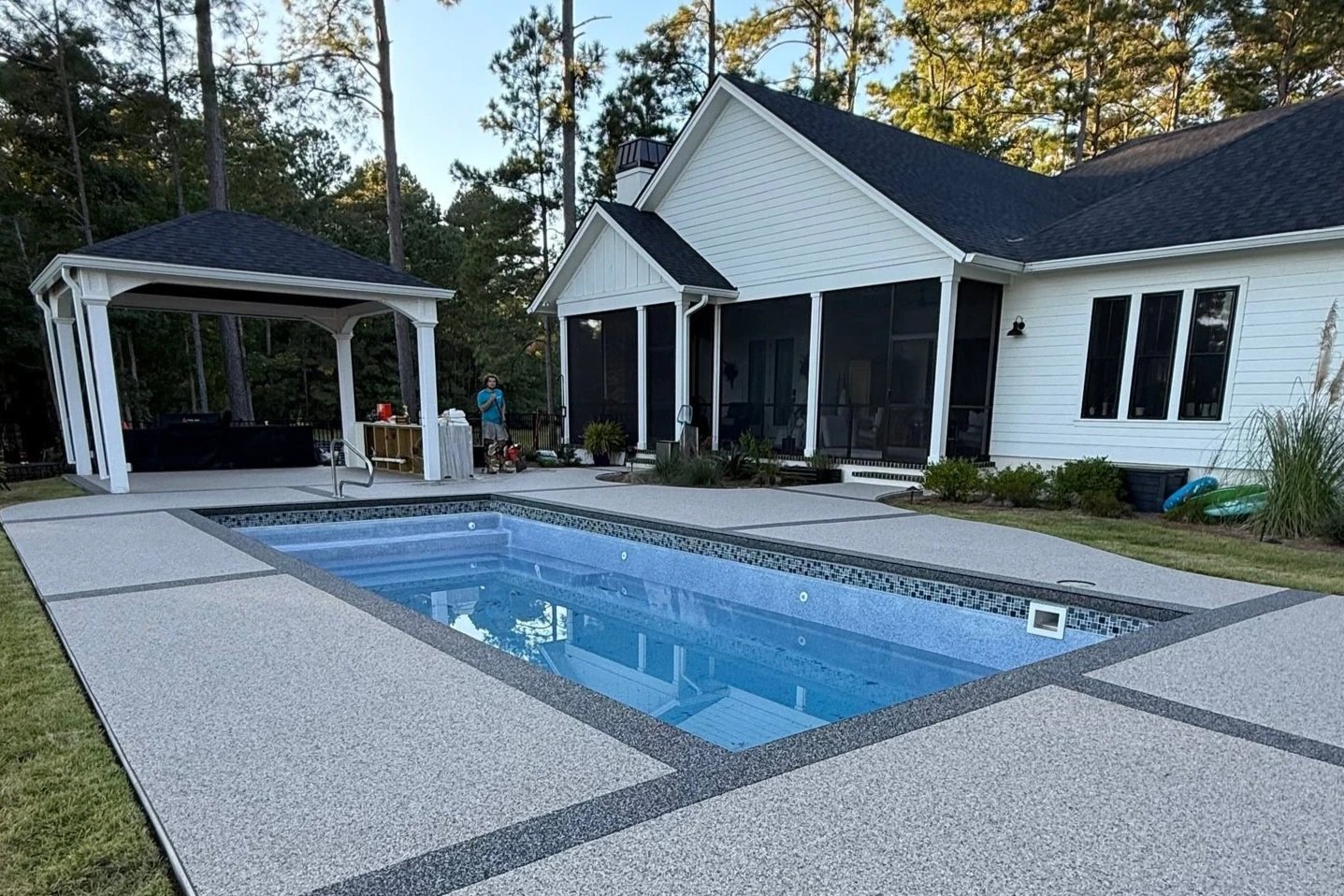Backyard with in-ground swimming pool surrounded by a concrete deck, there is a gazebo to the left with a hot tub inside, a person standing near a wooden table, and a white house with large windows and a screened porch in the background.