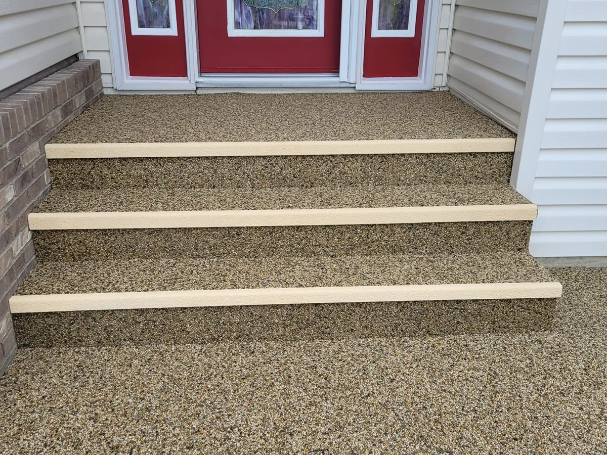Outdoor front steps with three steps, textured pebbled surface, with beige wooden boards as stair risers. Red front door with windows, white vinyl siding on house.