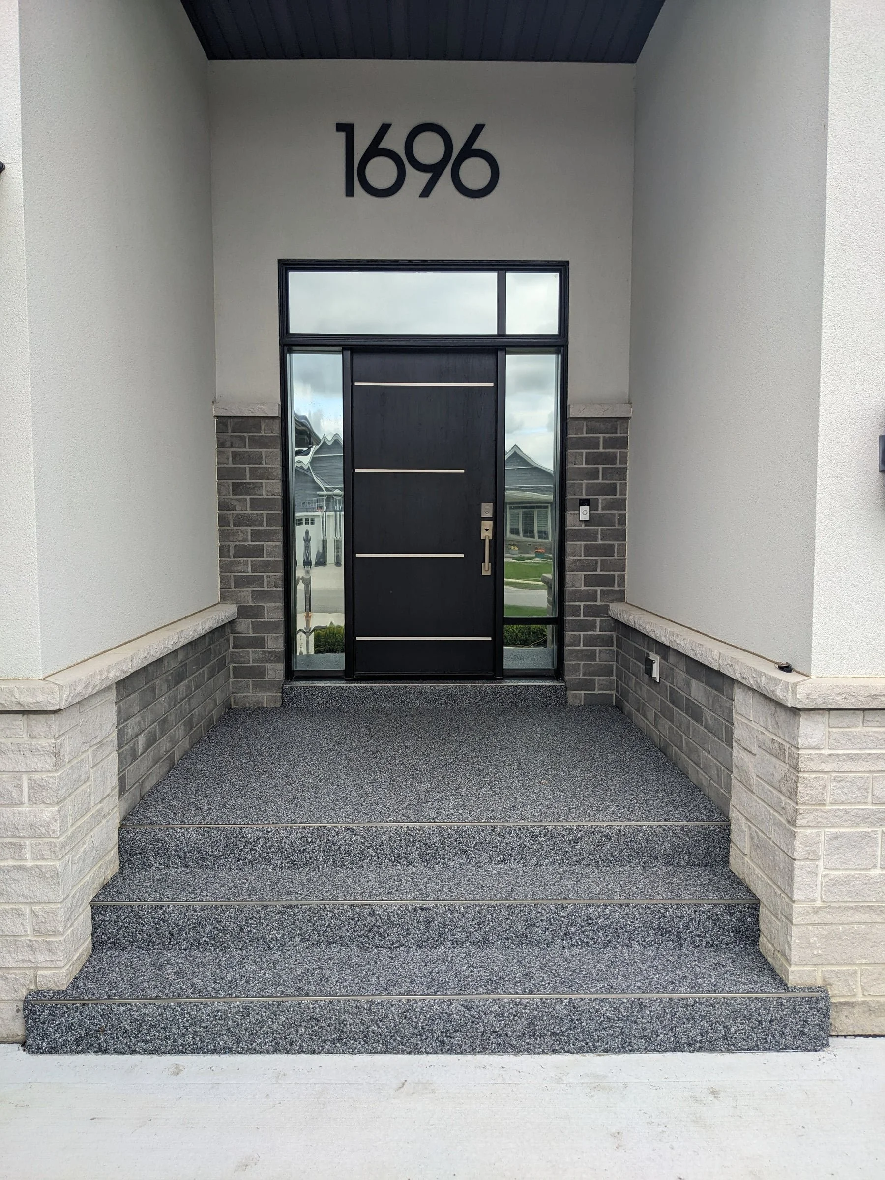 Front entrance of a house with a black door, gray brick accents, and the house number 1696 displayed above the door. The porch is covered, with a textured gray floor and cream-colored walls.