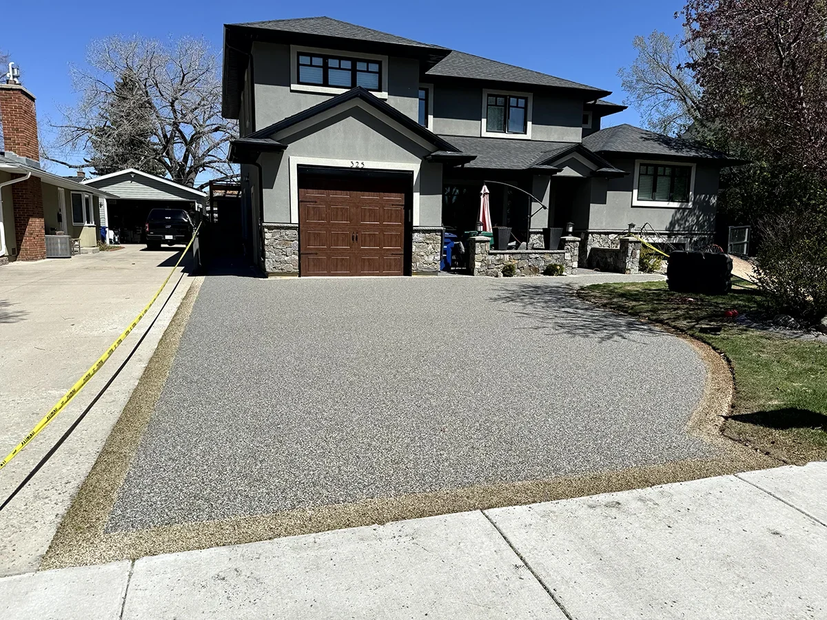A newly landscaped driveway in front of a two-story house with a gray exterior, stone accents, and a wooden garage door. The driveway is bordered with decorative gravel, and a yellow caution tape is stretched along one side.