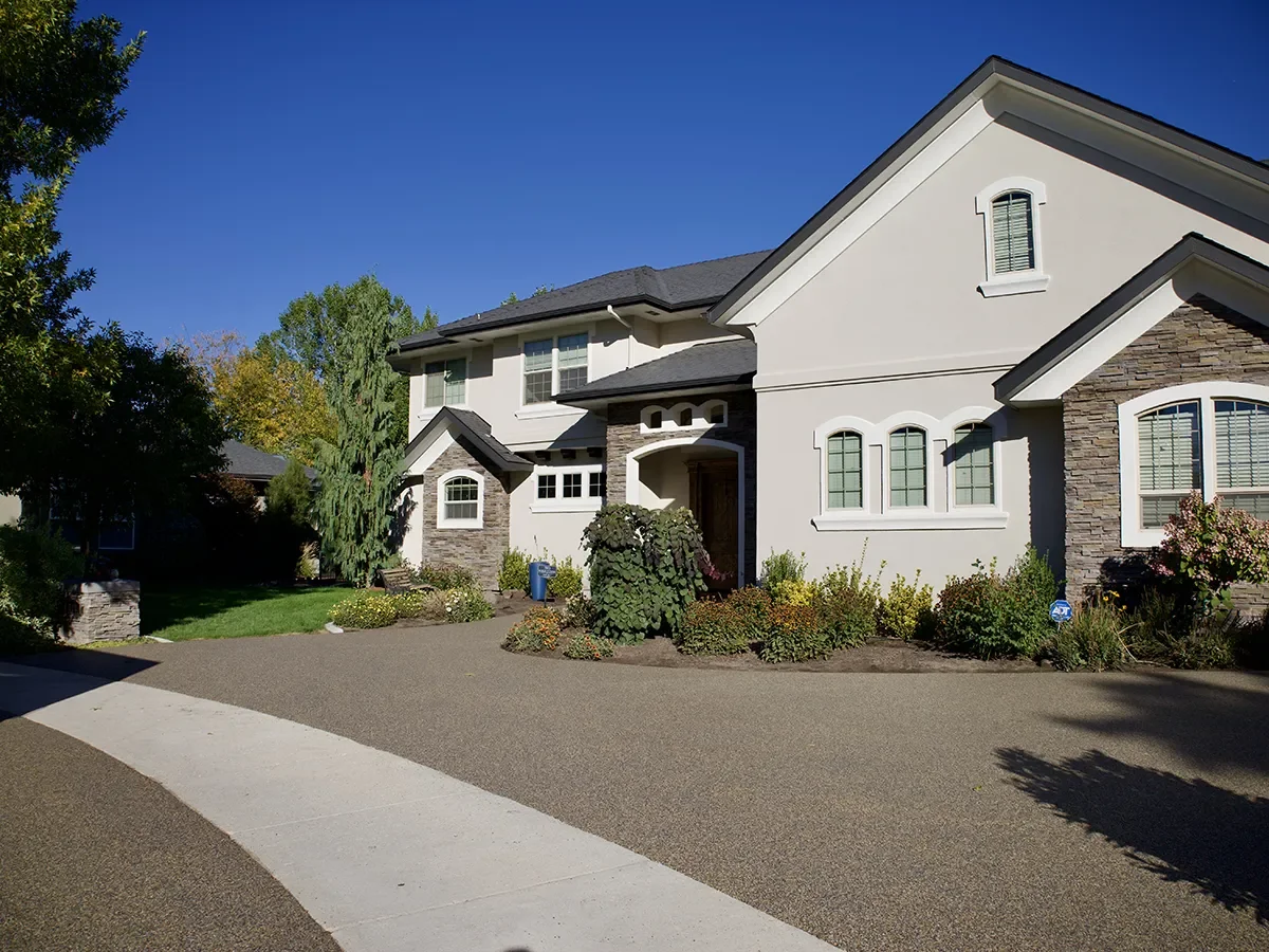 Front view of a modern two-story house with white walls, stone accents, and multiple windows, surrounded by landscaped garden and driveway under a clear blue sky.