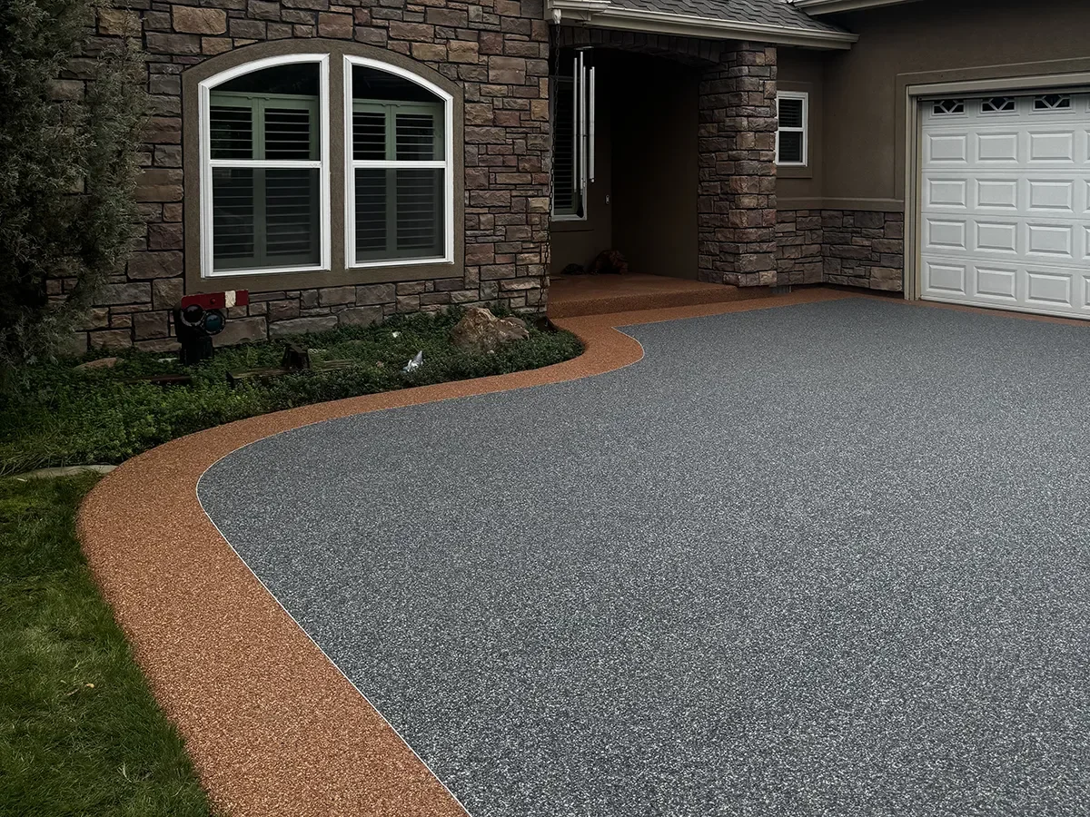 Concrete driveway with decorative border in front of a house with stone and siding exterior, large window with shutters, and a garage door.