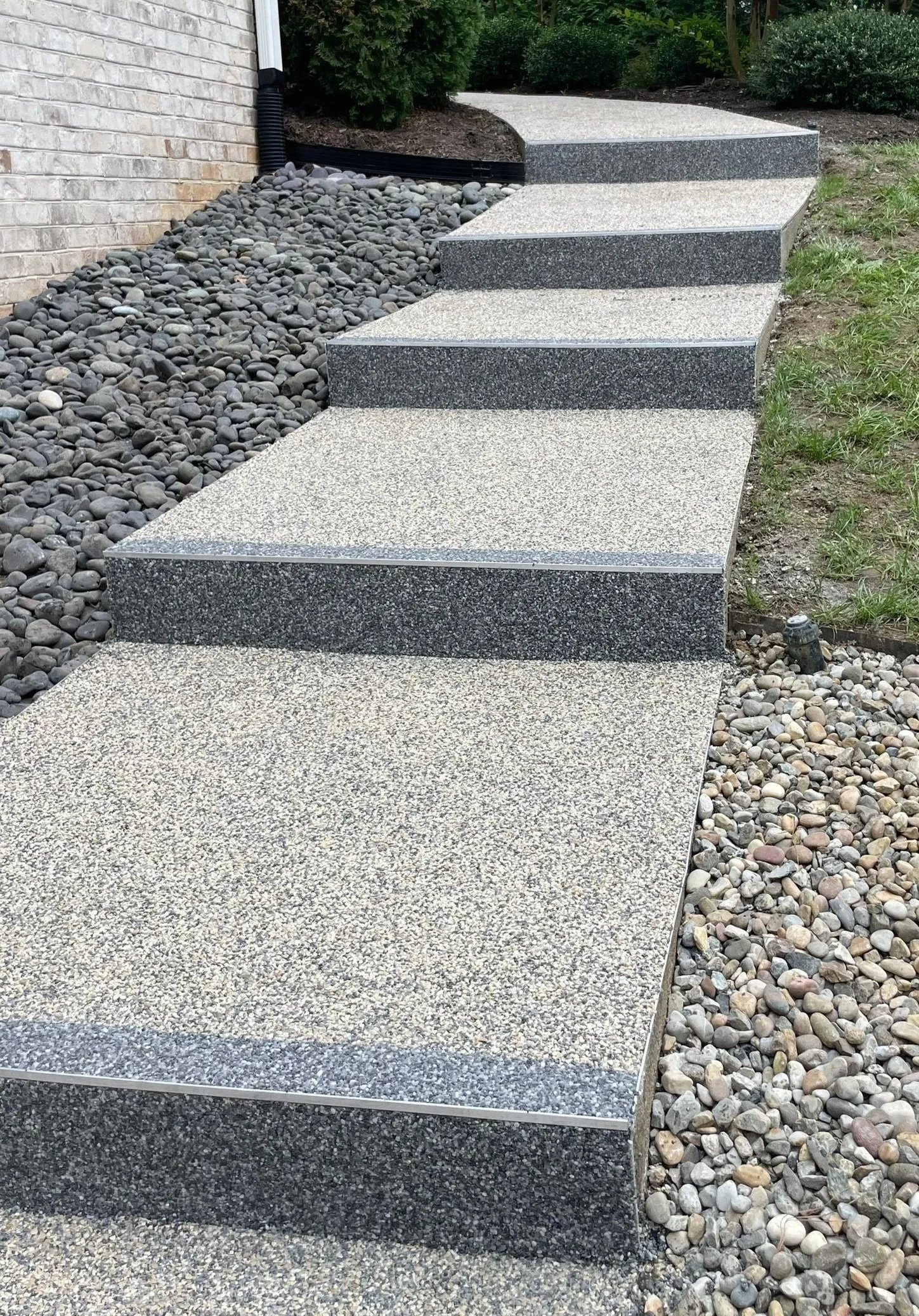 Concrete steps leading up to a house with rocks and small pebbles on the side, and a garden bed with bushes and grass.
