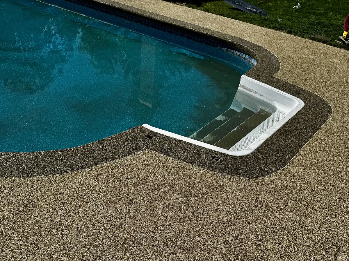 An empty swimming pool with a set of steps leading into the water, surrounded by a textured concrete deck, with some grass and outdoor equipment in the background.