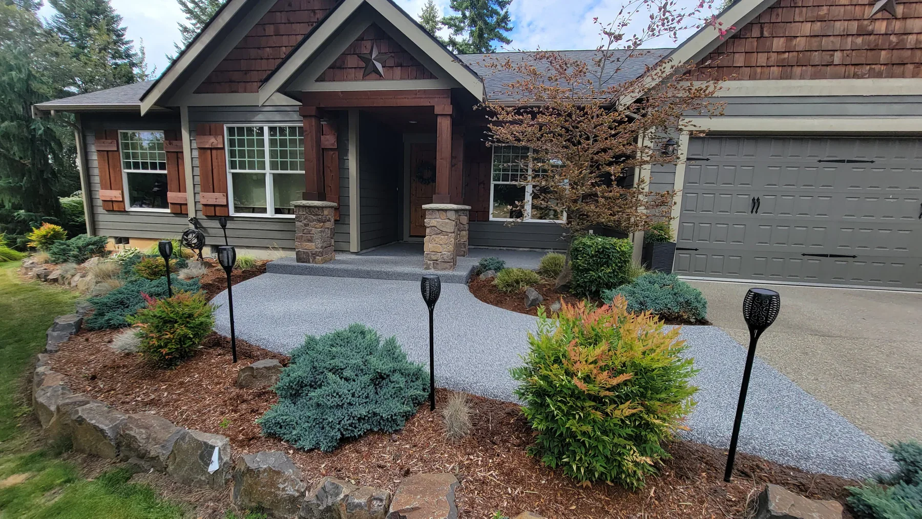 Front yard of a house featuring a stone paved walkway, garden beds with shrubs and a small tree, black decorative garden lights, and a gray garage door. The house has a porch with wooden columns and shutters, and the exterior combines gray siding wit