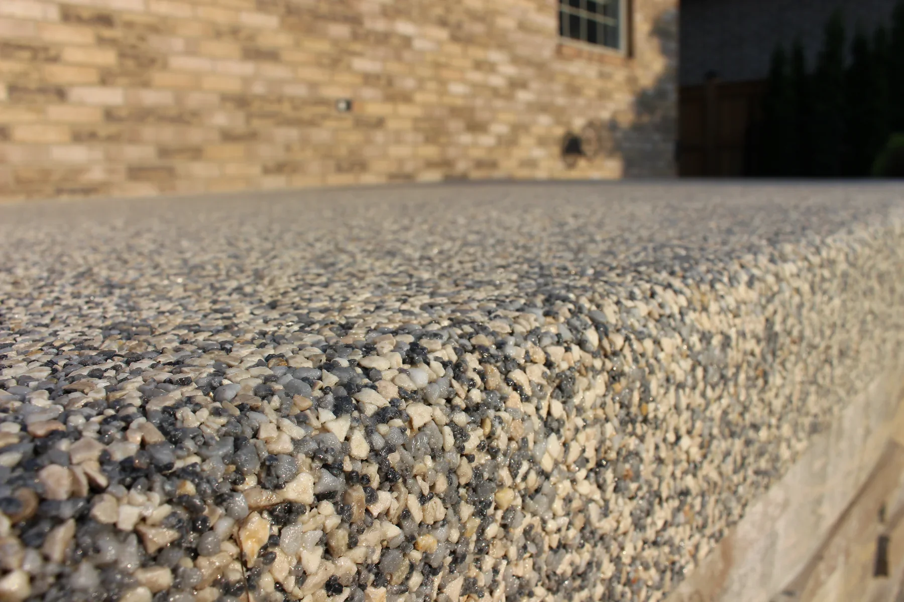 Close-up of a pebble-textured concrete surface with a brick house wall in the background.