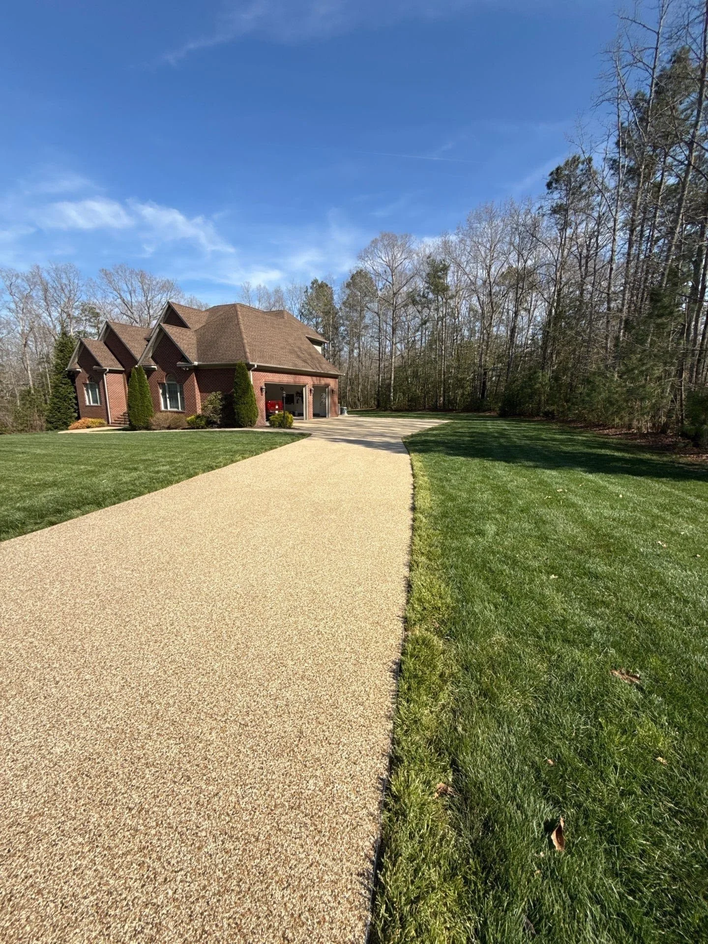 A brick house with an attached garage on a large lawn, with a paved driveway leading to the garage, surrounded by trees and a clear blue sky.