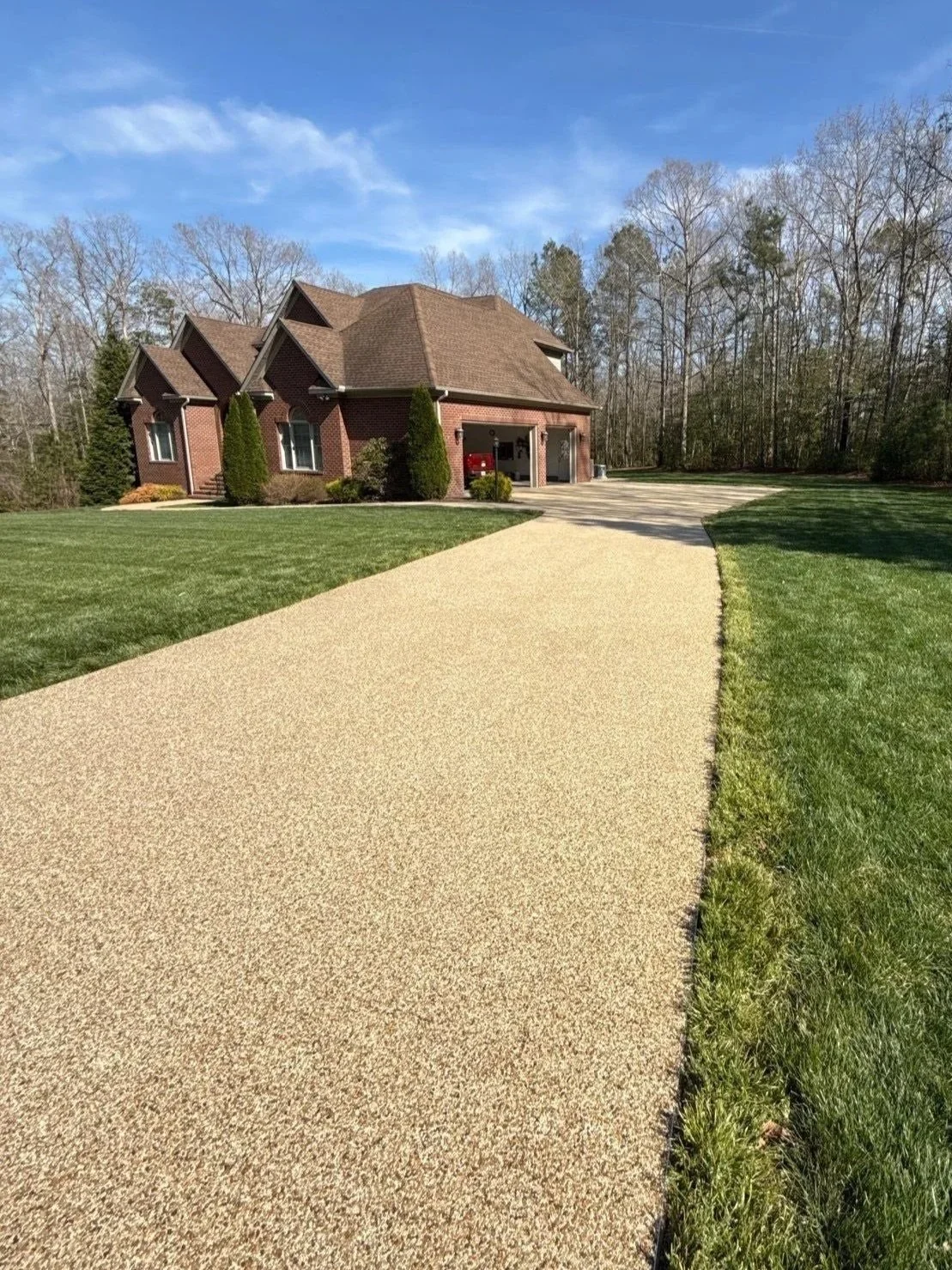 A brick house with a brown roof, surrounded by a well-maintained lawn and trees, with a textured concrete driveway leading to a garage on a clear day.