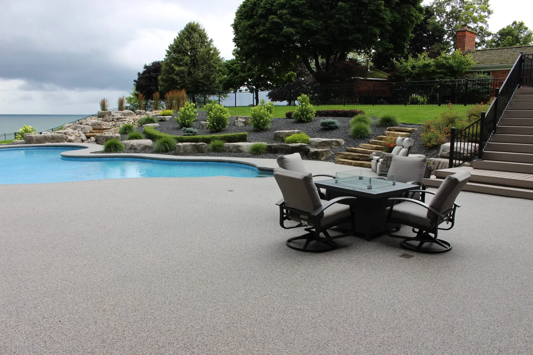 Backyard with swimming pool, outdoor furniture, landscaped garden, trees, and view of lake or ocean in the background under cloudy sky.