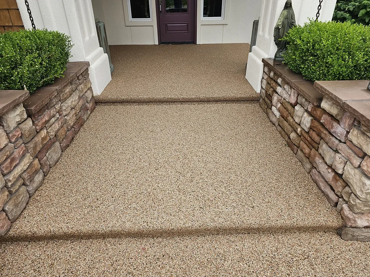 Front porch with textured beige outdoor carpet, stone side walls, green bushes, and a statue on the right side.