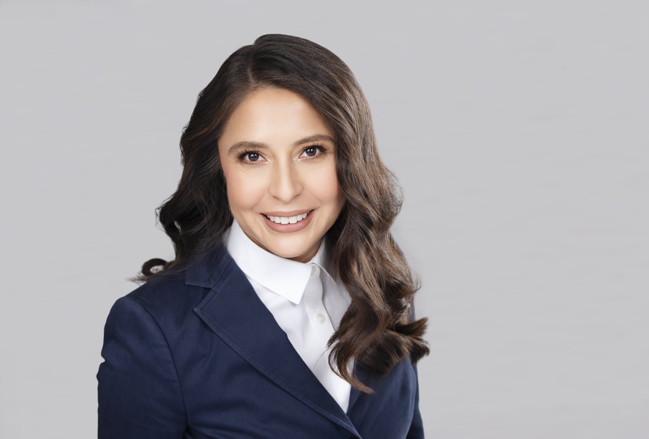 A woman with long wavy brown hair, wearing a navy blazer and white shirt, smiling against a plain gray background.