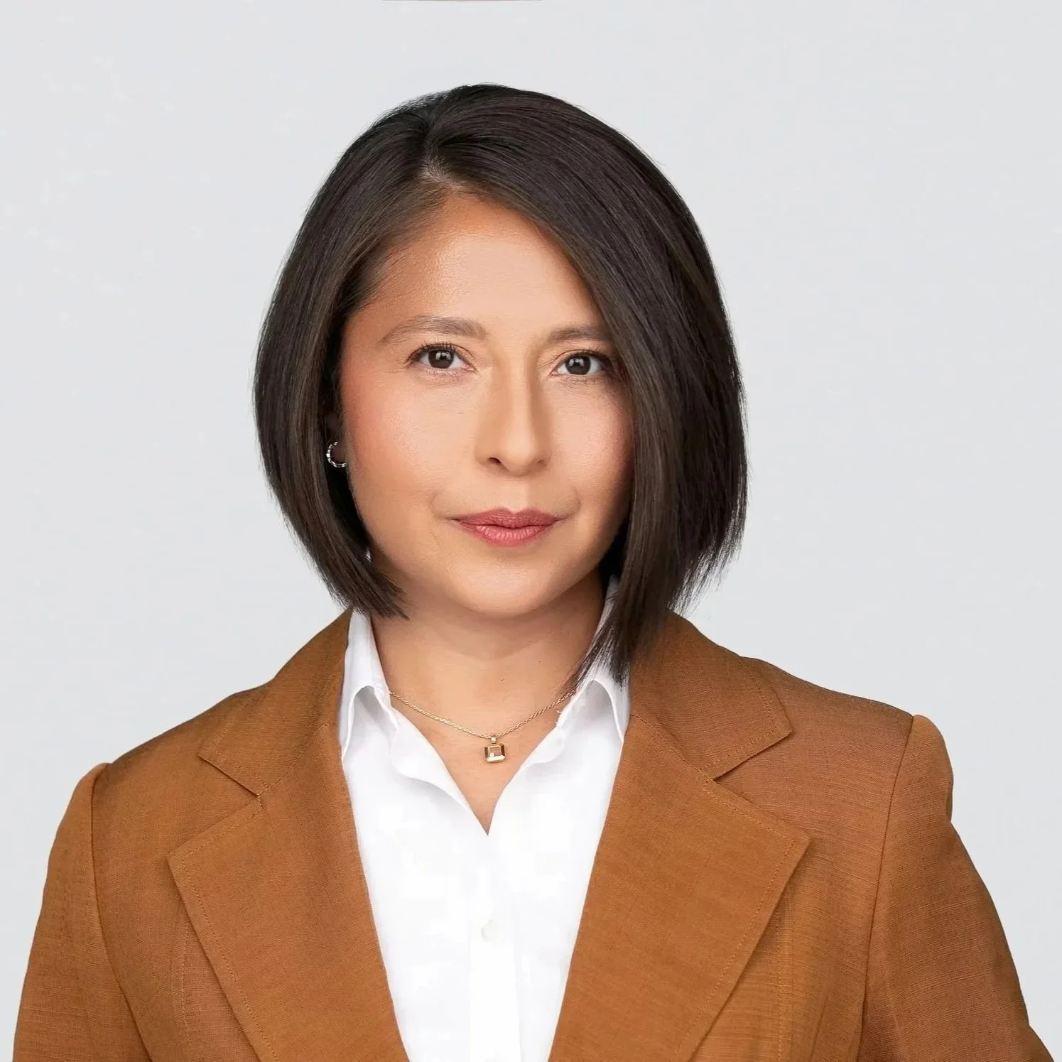 A woman with shoulder-length dark hair wearing a white shirt and a brown blazer, looking directly at the camera against a plain light gray background.