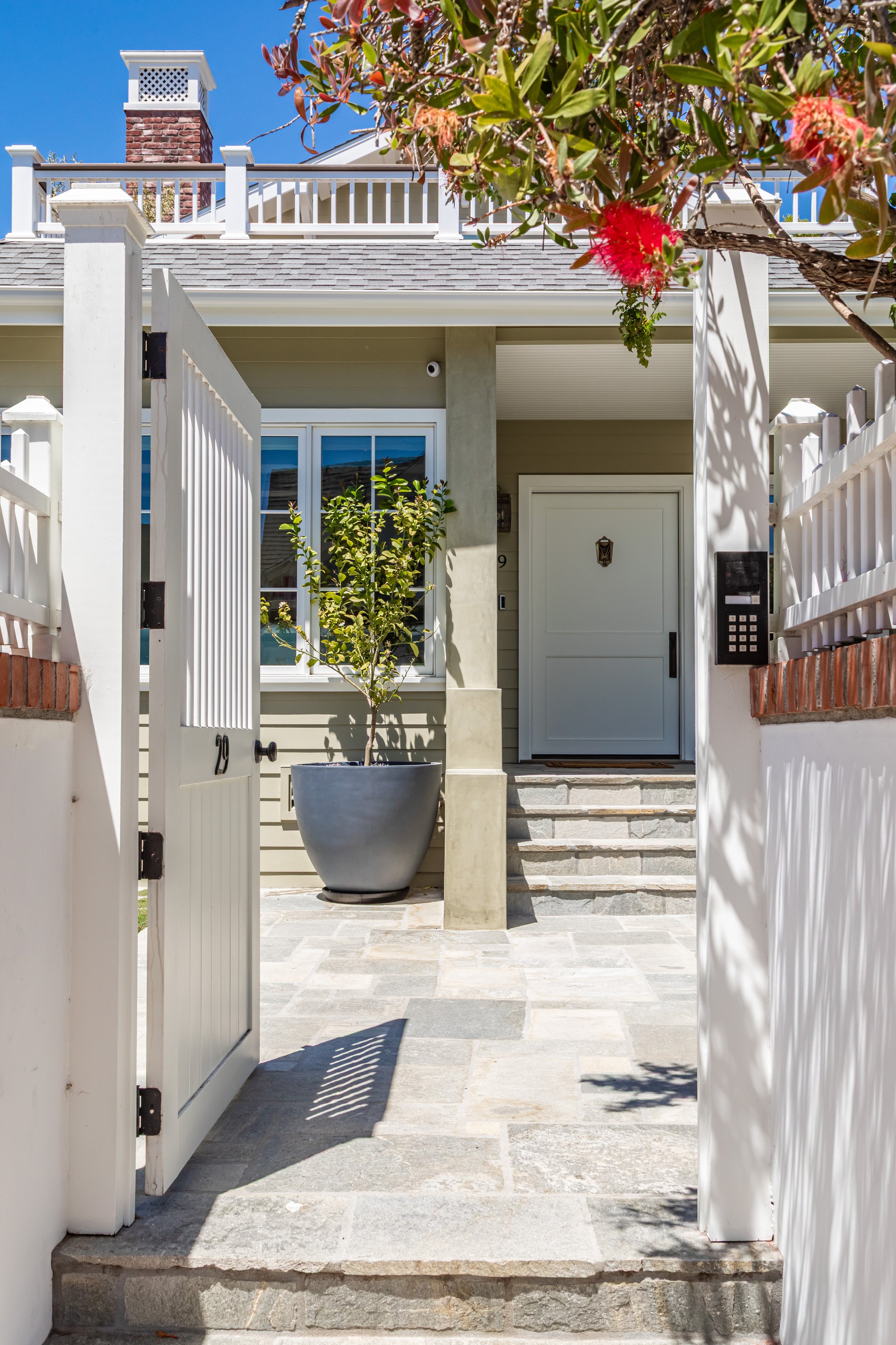 Front porch of a house with stairs, potted plant, and white gate under a blue sky.
