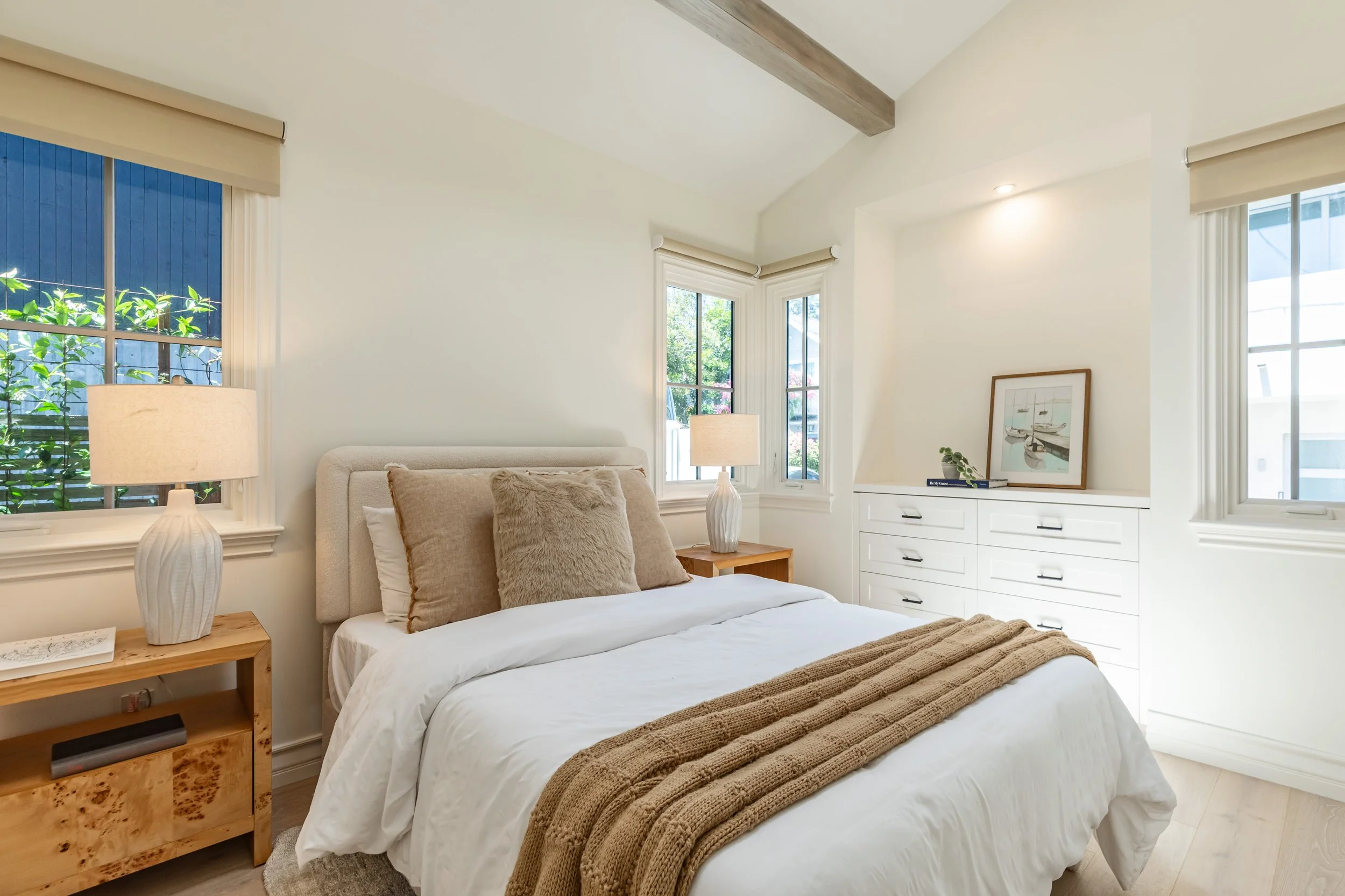 A bedroom with white walls, a large bed with white bedding and beige pillows, and two windows with cream-colored blinds. There are two white lamps on wooden nightstands on either side of the bed, and a white dresser with drawers and a framed picture 