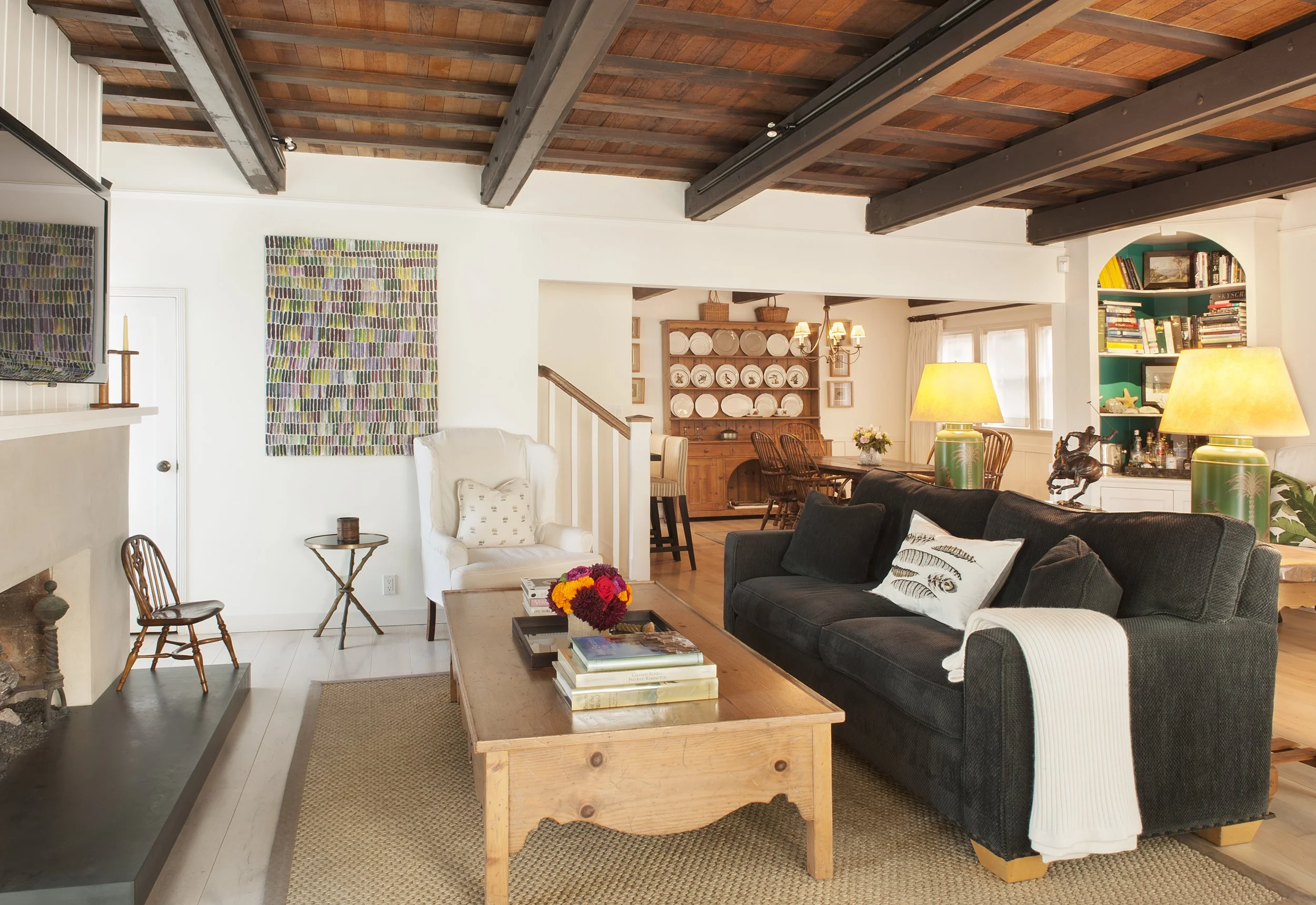 Living room with white walls, a wooden ceiling with exposed beams, a dark sofa, a white armchair, a coffee table with books and a flower arrangement, a fireplace, a wooden dining table with chairs, a bookshelf, and artwork on the walls.