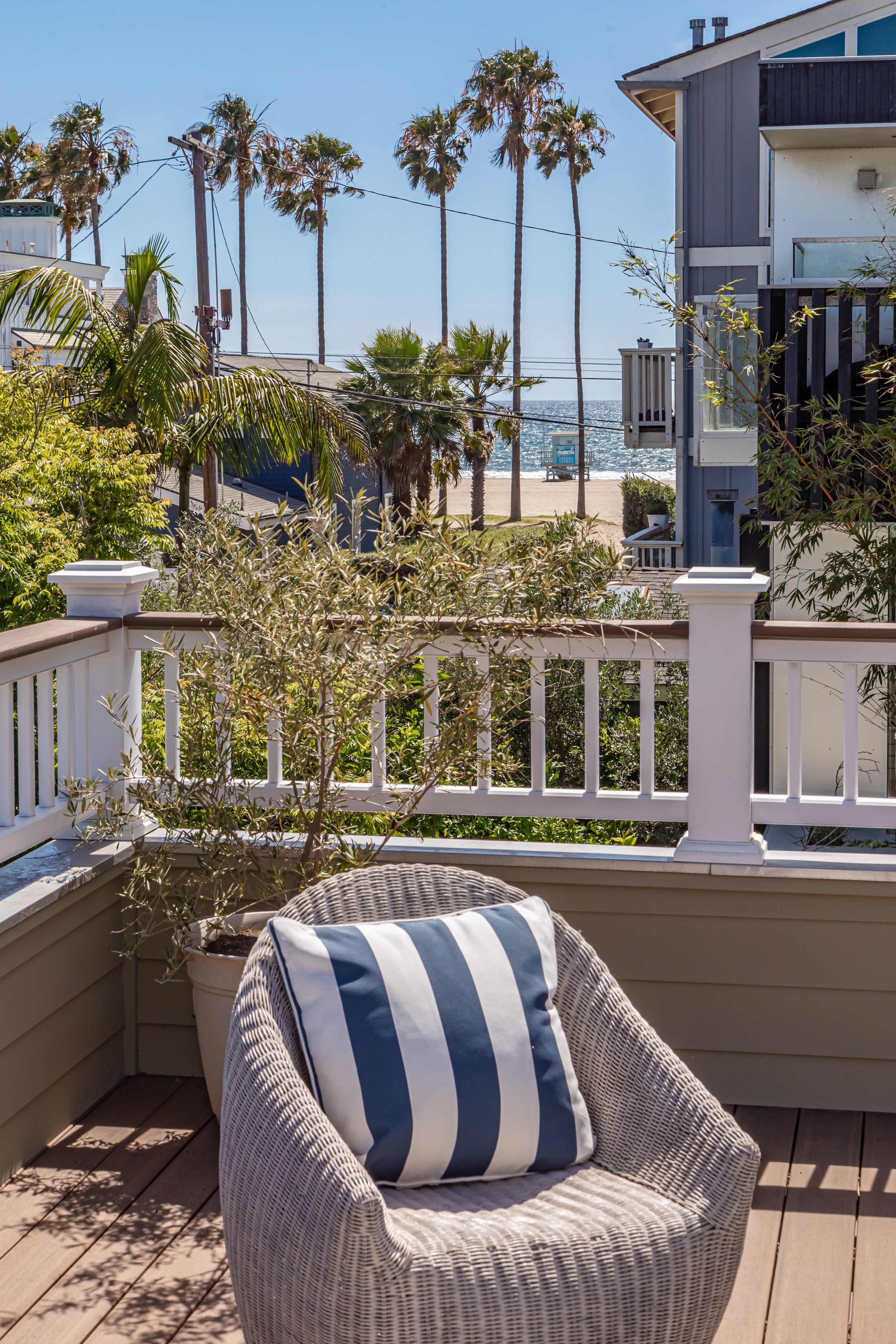 A balcony with a wicker chair and a striped cushion, surrounded by greenery, overlooking palm trees and a beach with the ocean in the background on a sunny day.