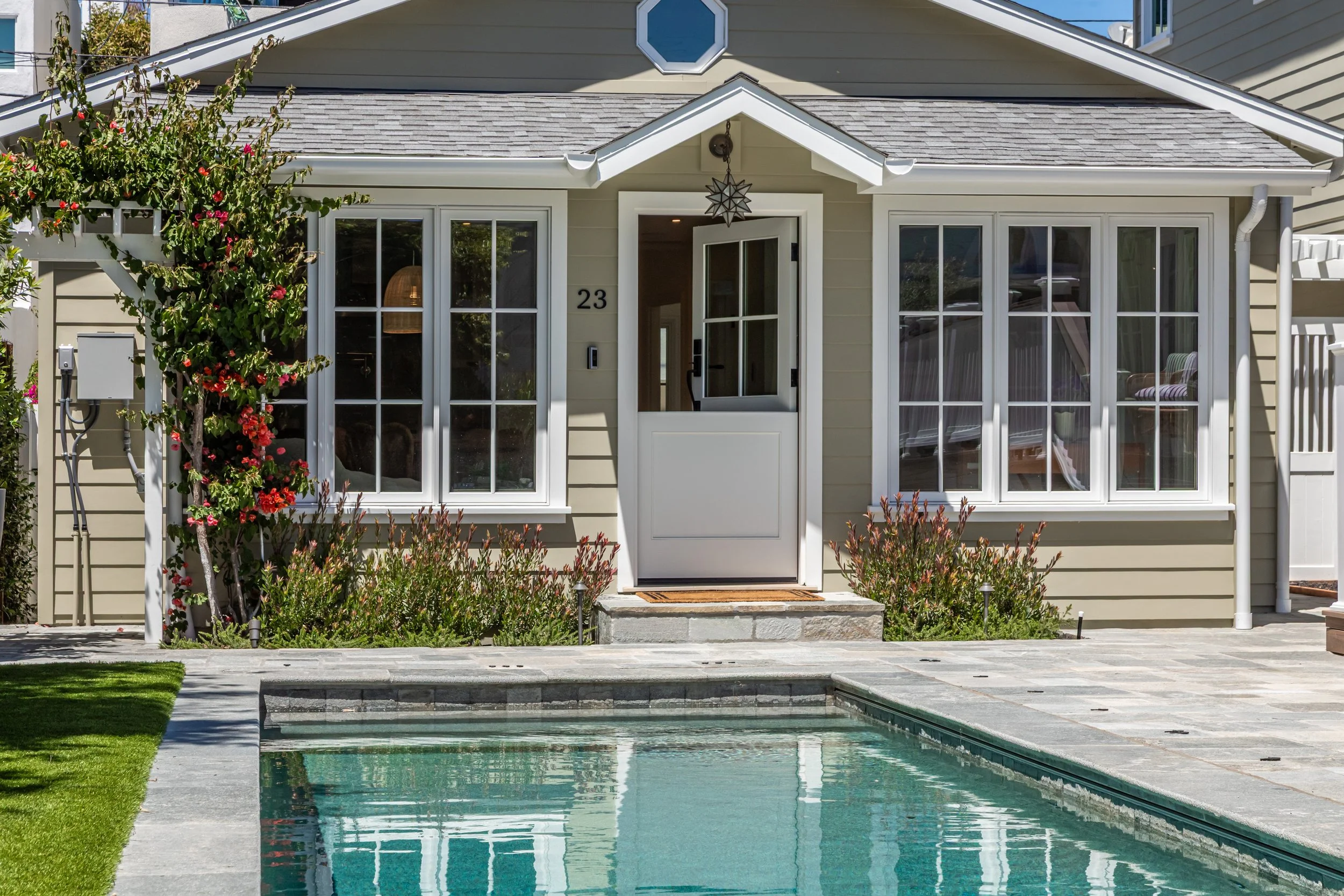 A backyard with a swimming pool, a beige house with large windows and a door, a small front yard with grass and bushes, and a flowering tree on the left side.