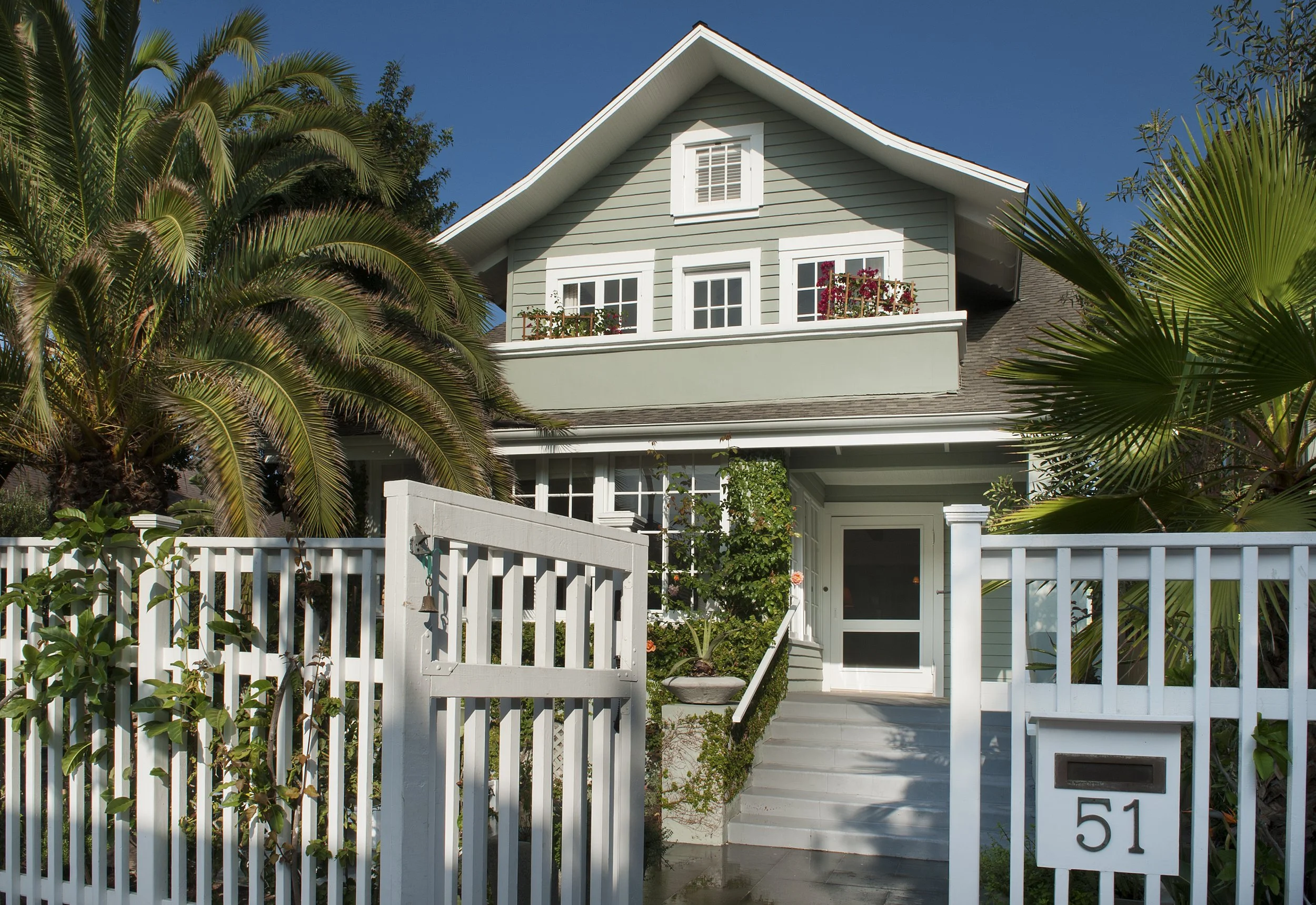 Two-story house painted light green with white trim, a small balcony with flowers, and a white picket fence with greenery and plants in the yard, under a clear blue sky.