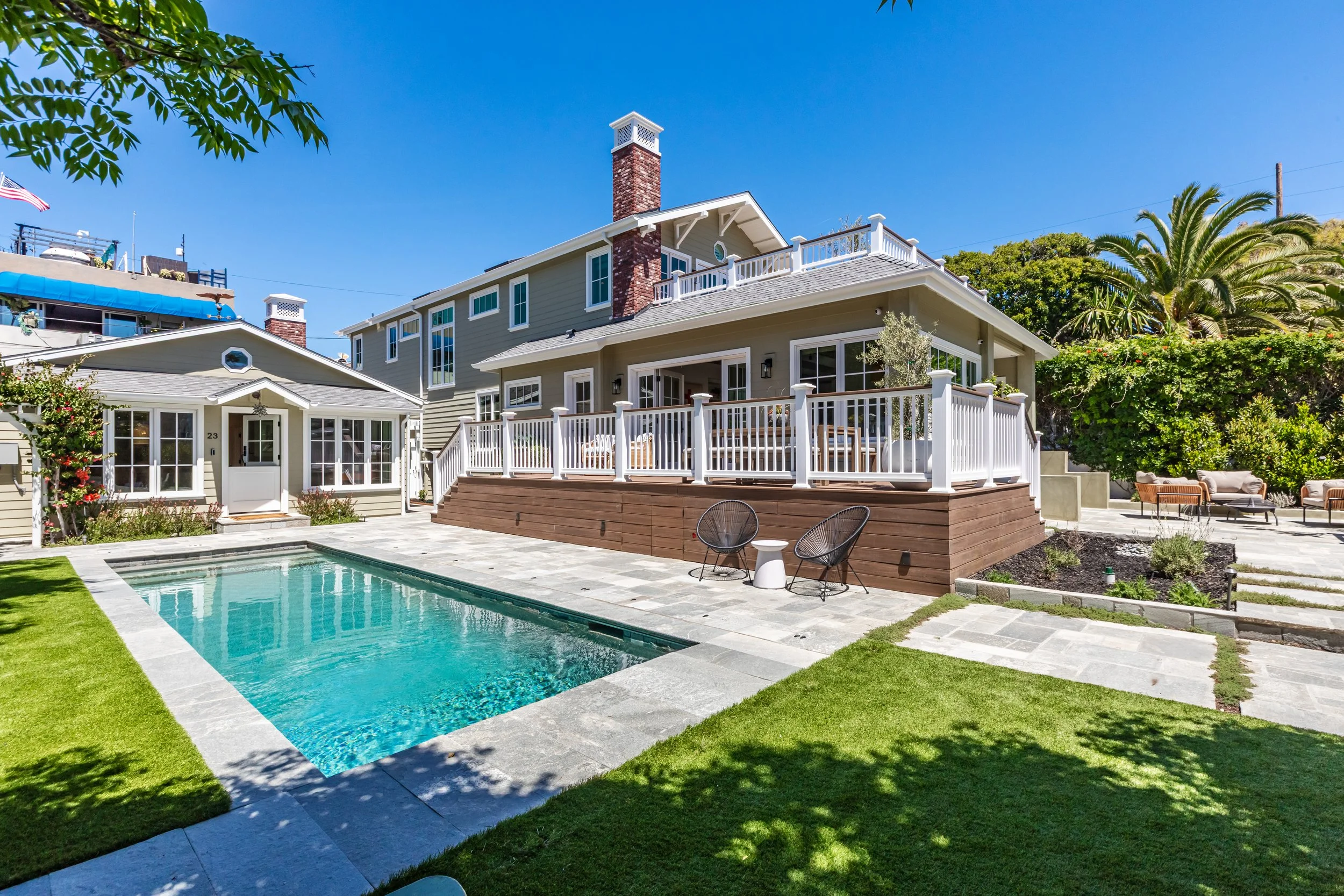 A backyard with a swimming pool, a wooden deck, and patio furniture, surrounded by a well-maintained lawn and greenery, with a multi-story house in the background under a clear blue sky.