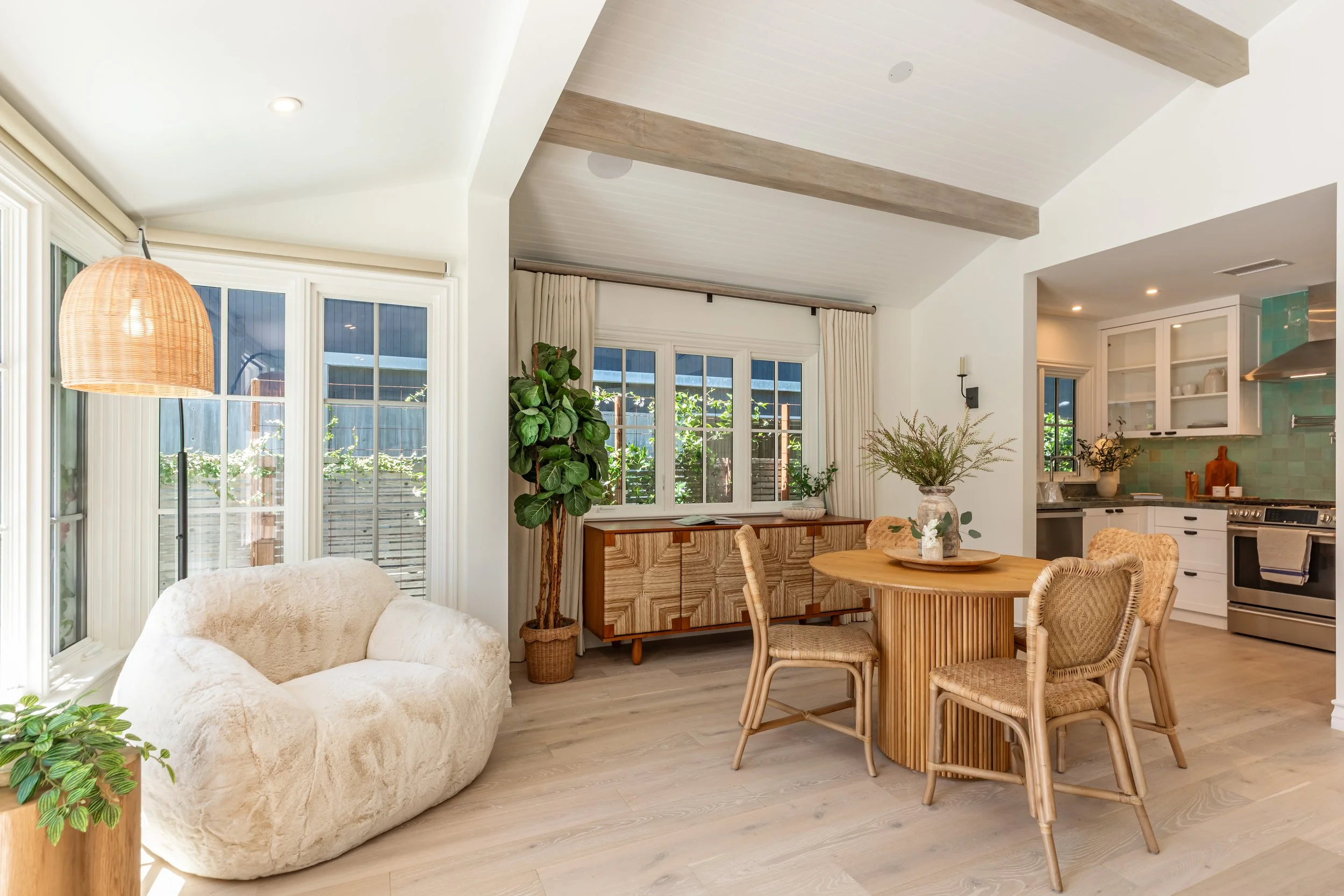 A bright, airy living and dining area with large windows, a cream-colored sofa, a round wooden dining table with wicker chairs, potted plants, and a view into the kitchen.