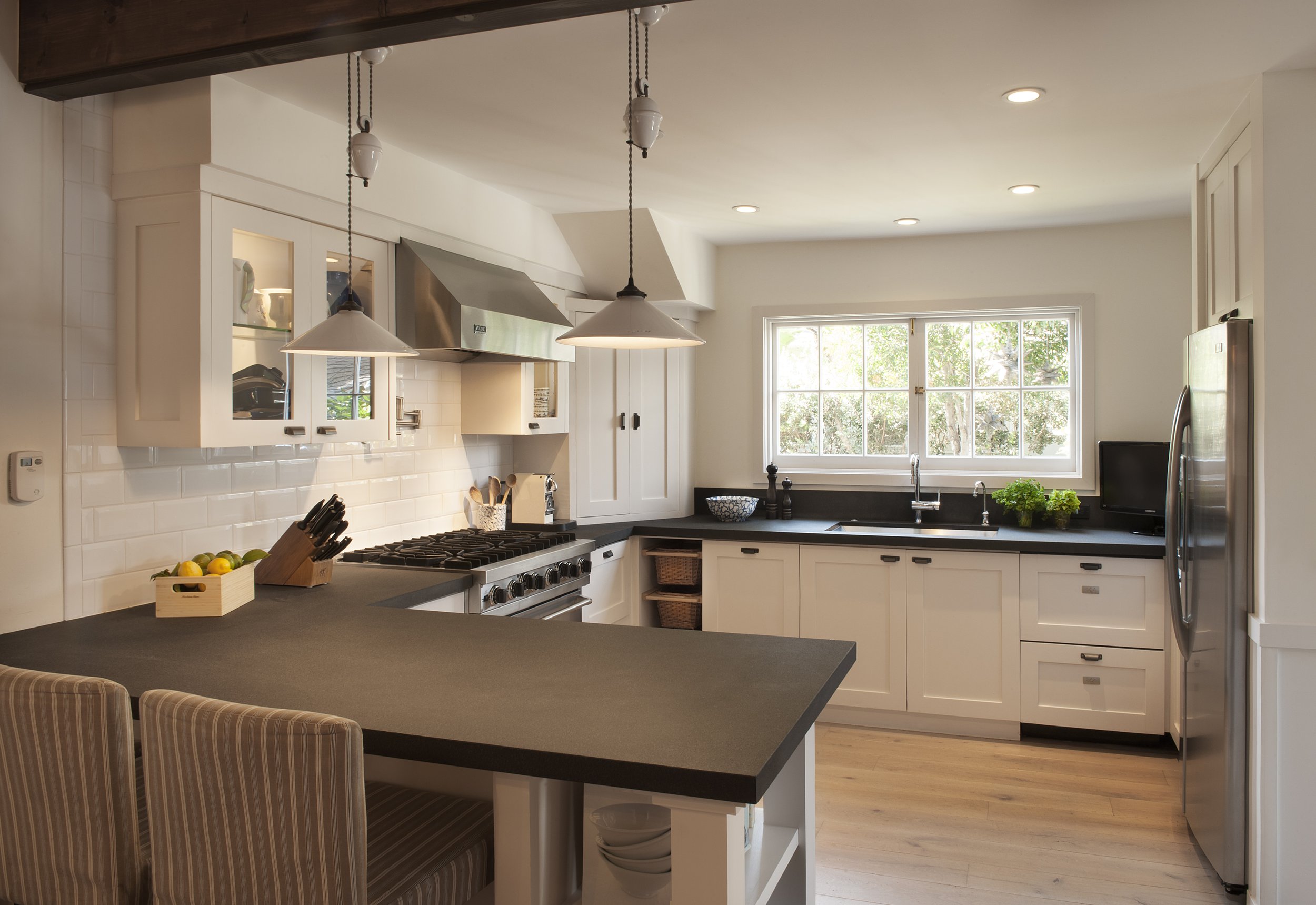 A modern, bright kitchen with white cabinets, stainless steel appliances, and a large window letting in natural light. There is a black countertop, hanging pendant lights, and a wooden basket with lemons on the counter.