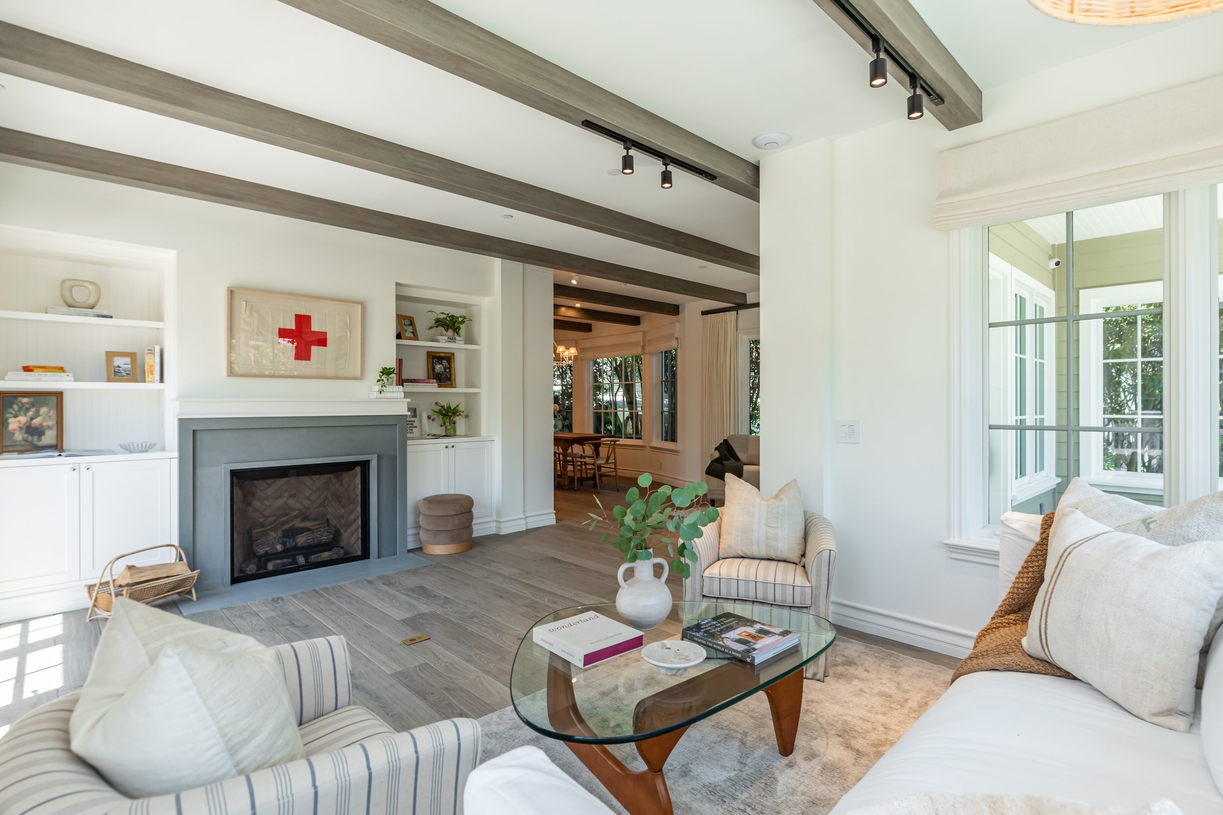 Bright living room with white walls and large windows. There is a fireplace with a gray mantel, built-in white shelves with decorations, and a beige armchair. A glass coffee table with books and a decorative tray is in front of a striped sofa and a w