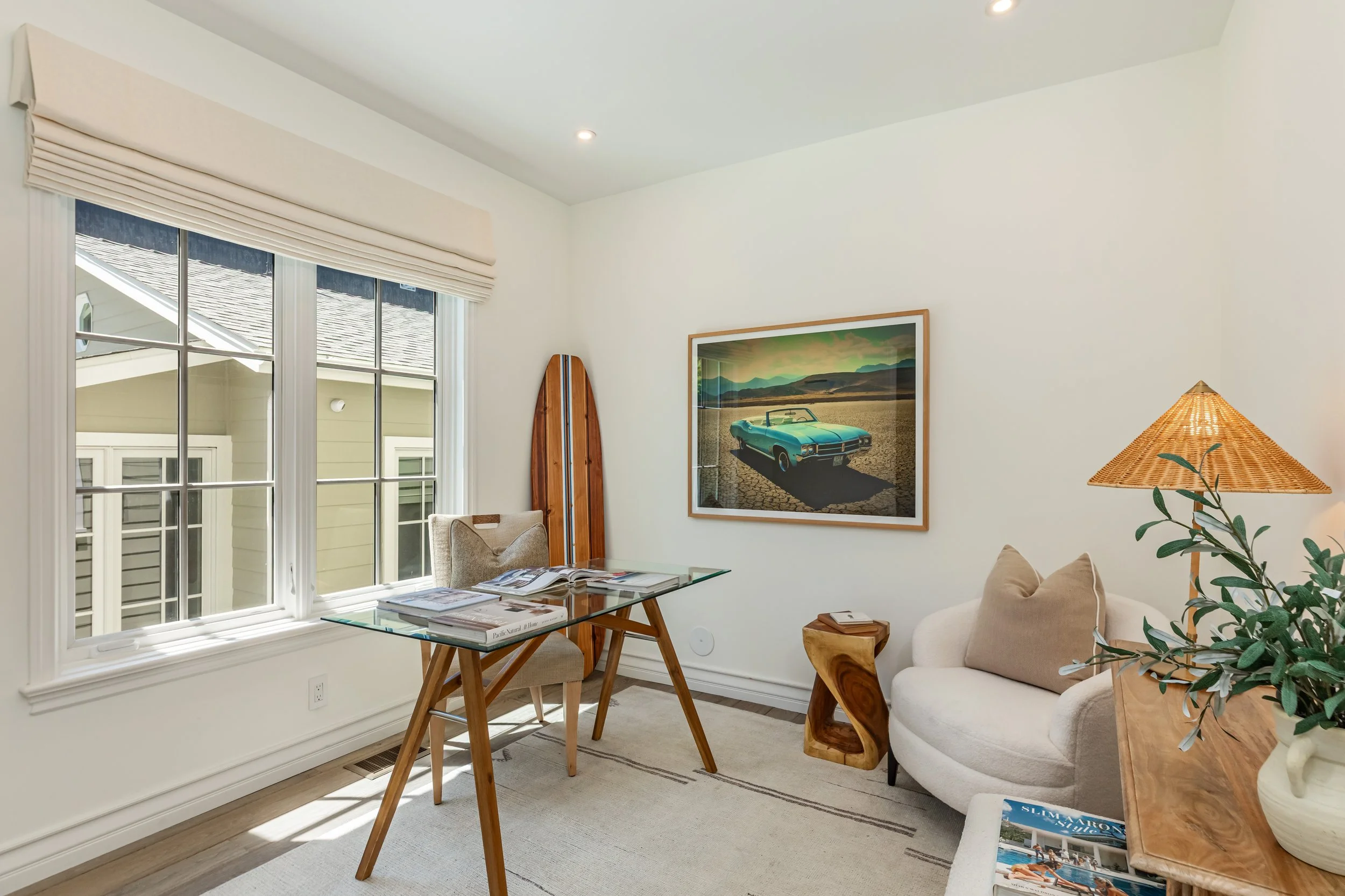 A cozy living room with large window, framed picture of a vintage car, wood and rattan furnishings, a table with magazines, and a tall houseplant.