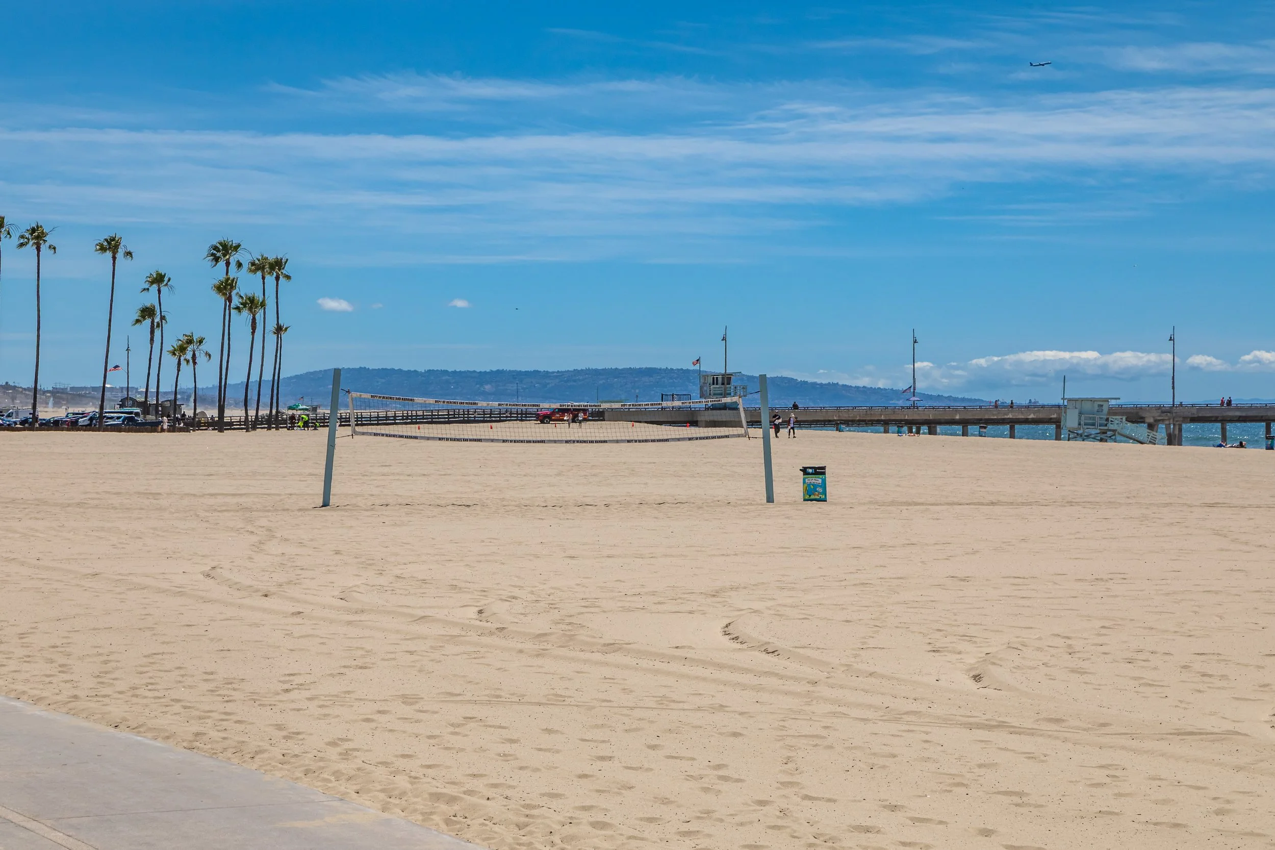 Sunny beach scene with volleyball court, palm trees, pier with people, and a distant airplane in the sky.