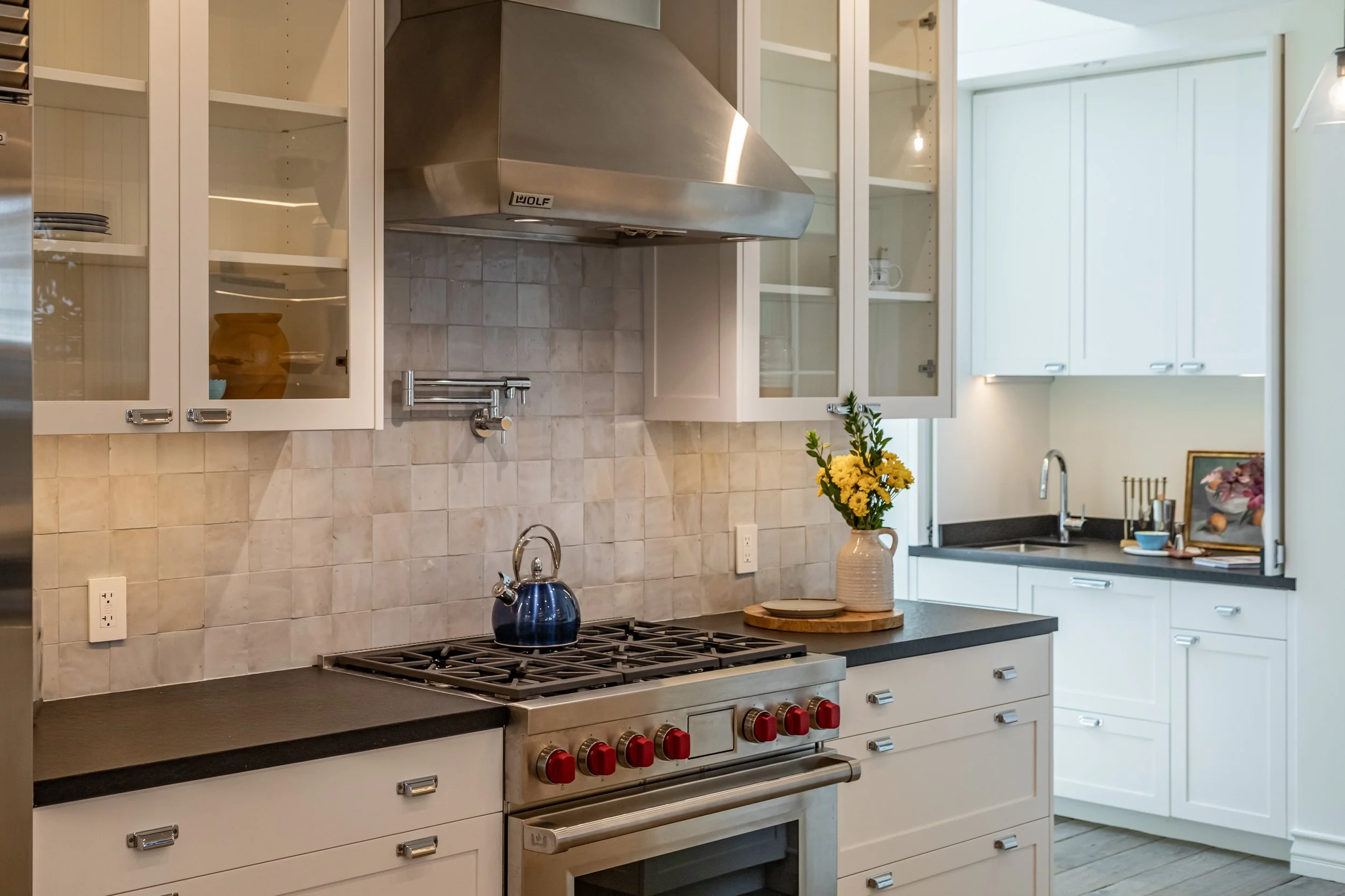 A kitchen with white cabinets, a black countertop, a stainless steel stove with red knobs, and a stove hood labeled 'Wolf'. There is a beige tiled backsplash, a blue kettle on the stovetop, and a vase with yellow flowers on the counter. To the right,