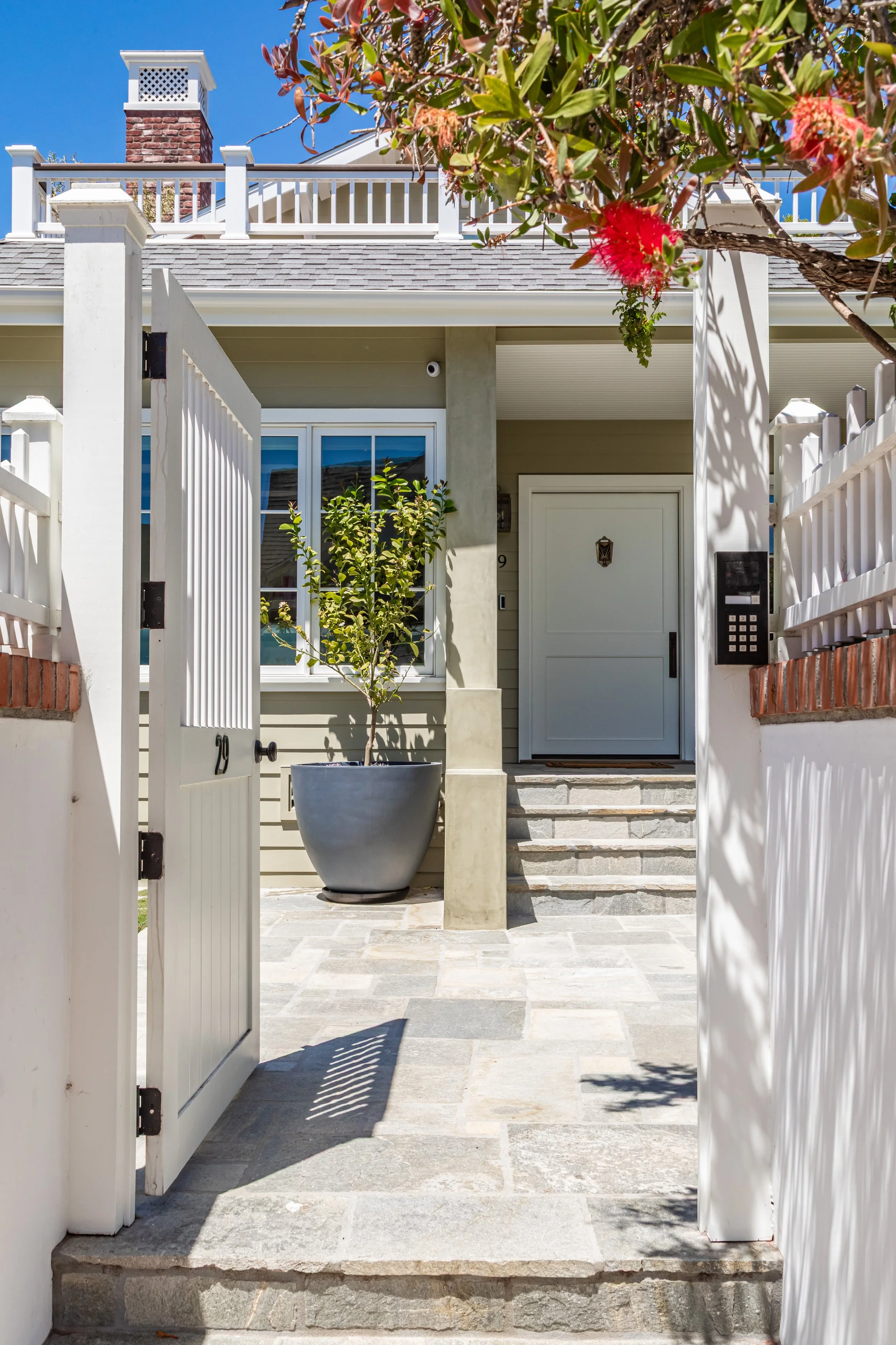 Front entrance of a house with open white gate, potted plant, steps leading to the front door, and a tree with red flowers in the foreground.