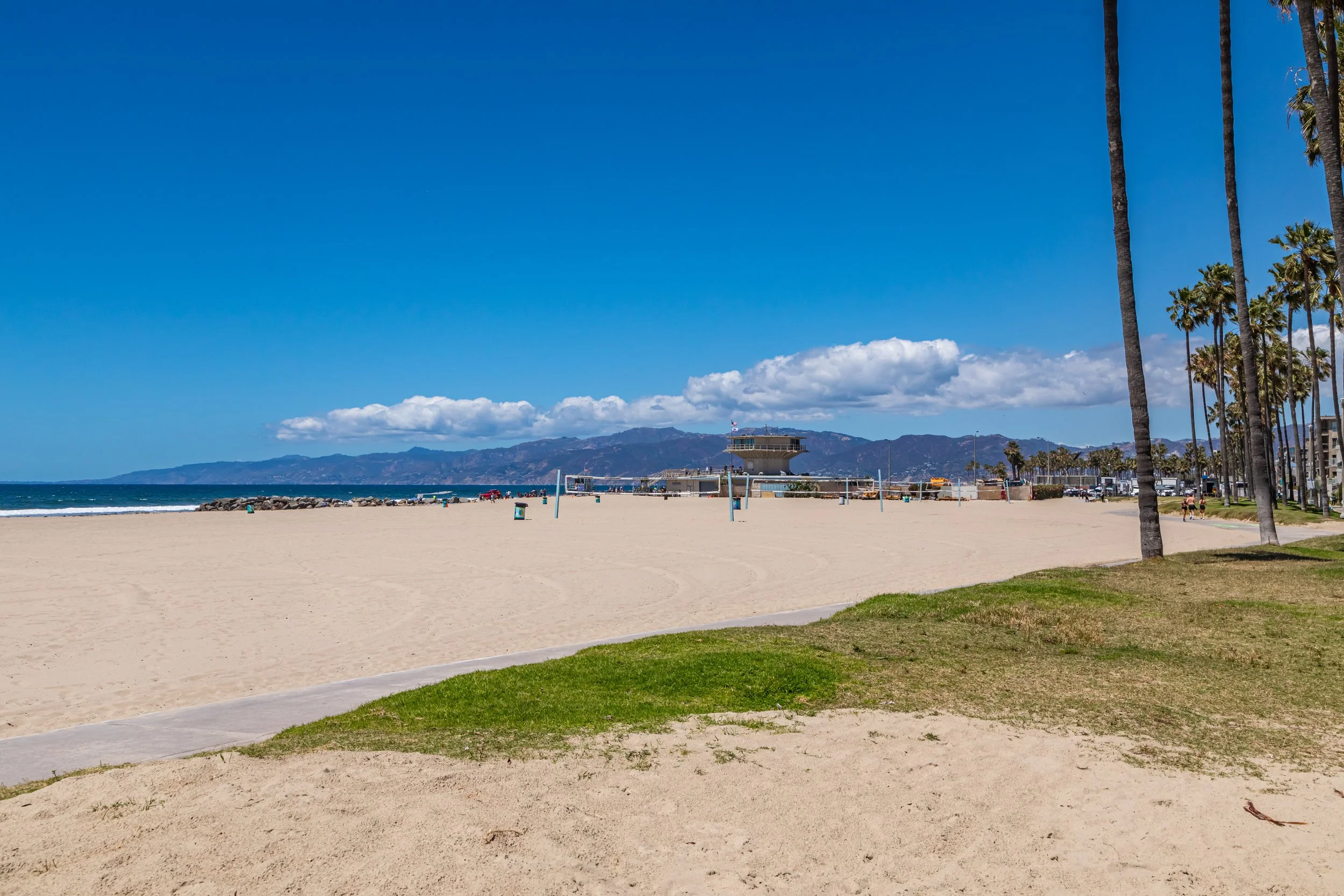 Sunny beach with sandy shore, ocean waves, palm trees, a lifeguard tower, and distant mountains under a blue sky with some clouds.