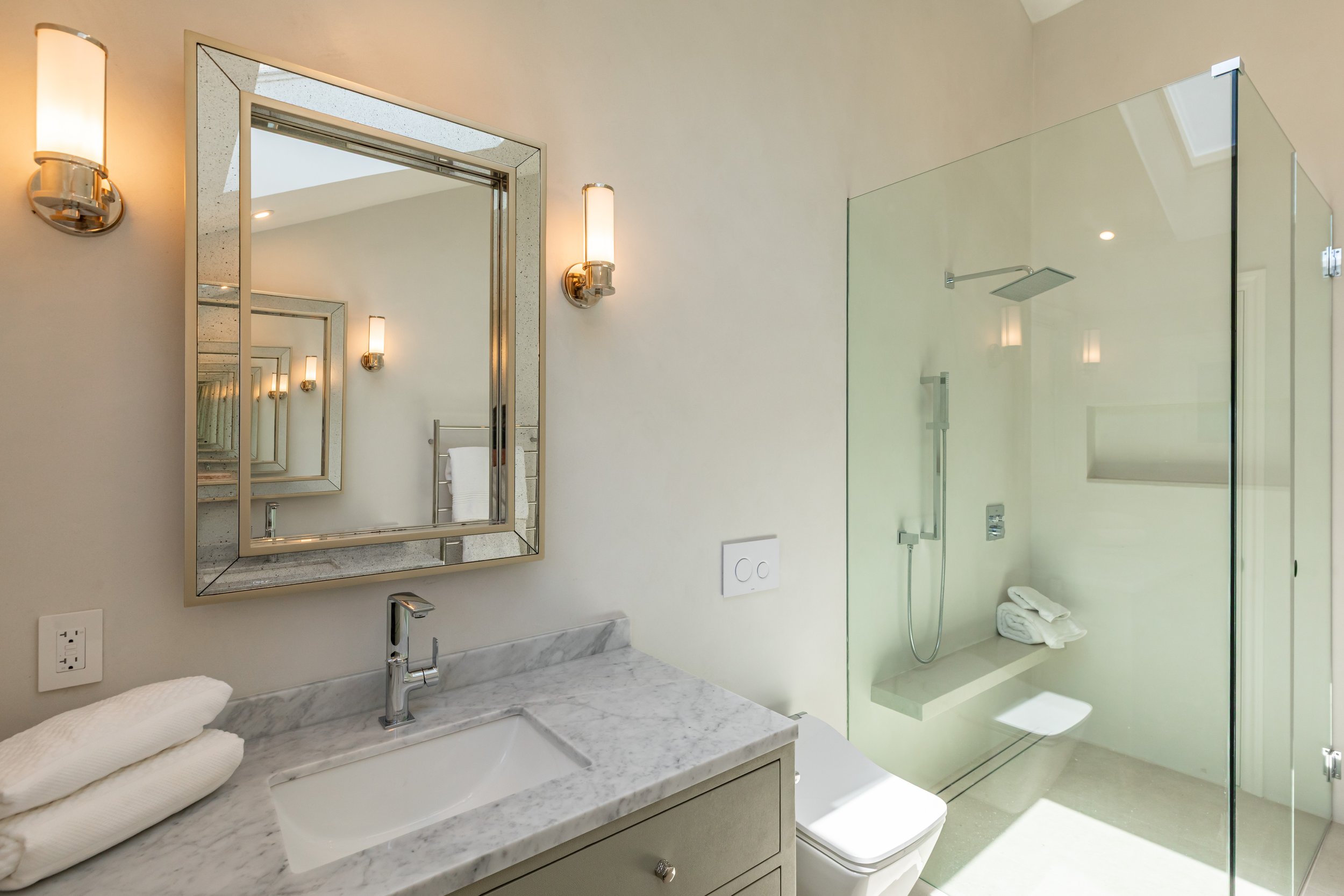 Bathroom with a marble countertop, a sink, a mirror, wall-mounted lights, a glass shower enclosure with a showerhead, and towels.