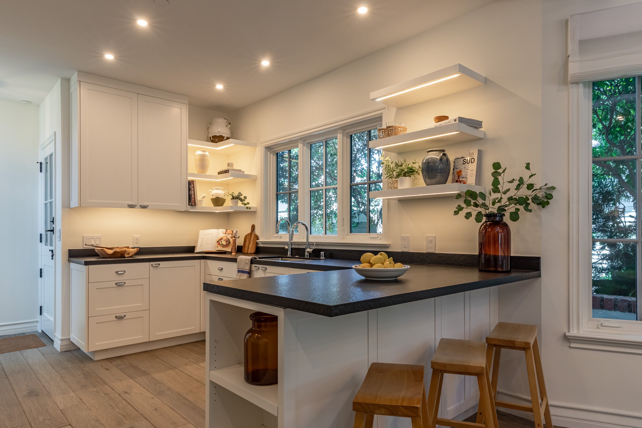 A modern kitchen with white cabinets, black countertops, a white island, and natural light from two large windows. Decor includes vases, a plant, books, and a bowl of lemons.