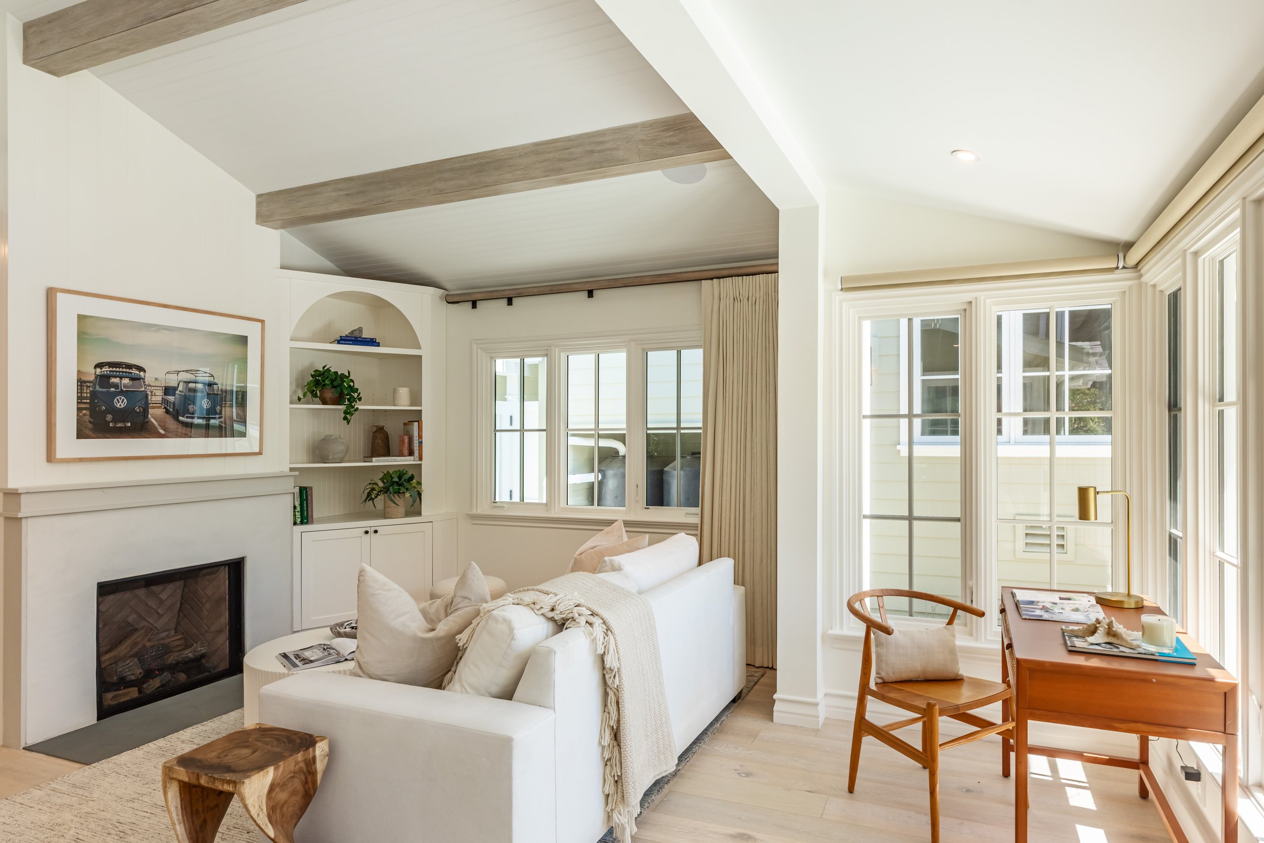 A bright living room with large windows, beige curtains, white walls, a white sofa with pillows, a wooden dining table with chairs, and decorative items on a built-in shelf, with exposed wooden beams on the ceiling.